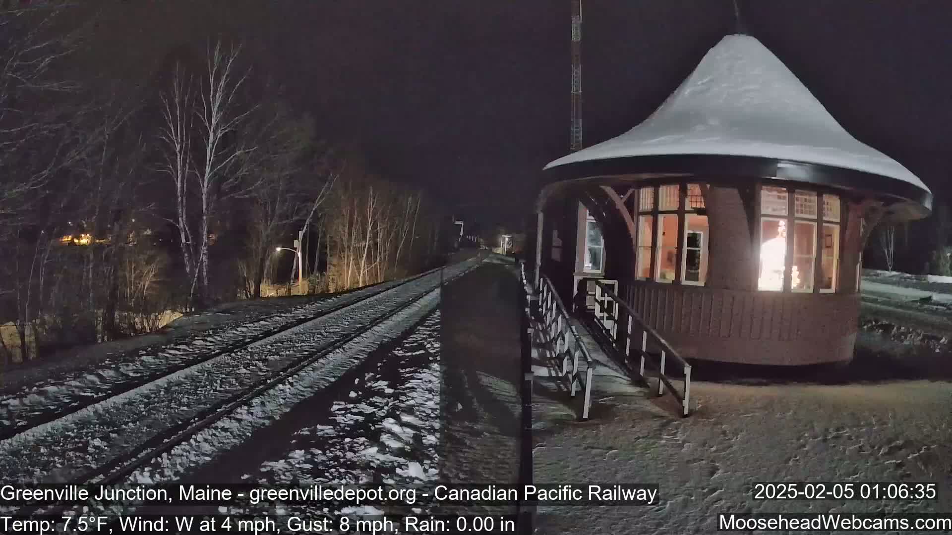 A snow-covered train depot with a lit interior is situated beside a snow-dusted railway track at night.