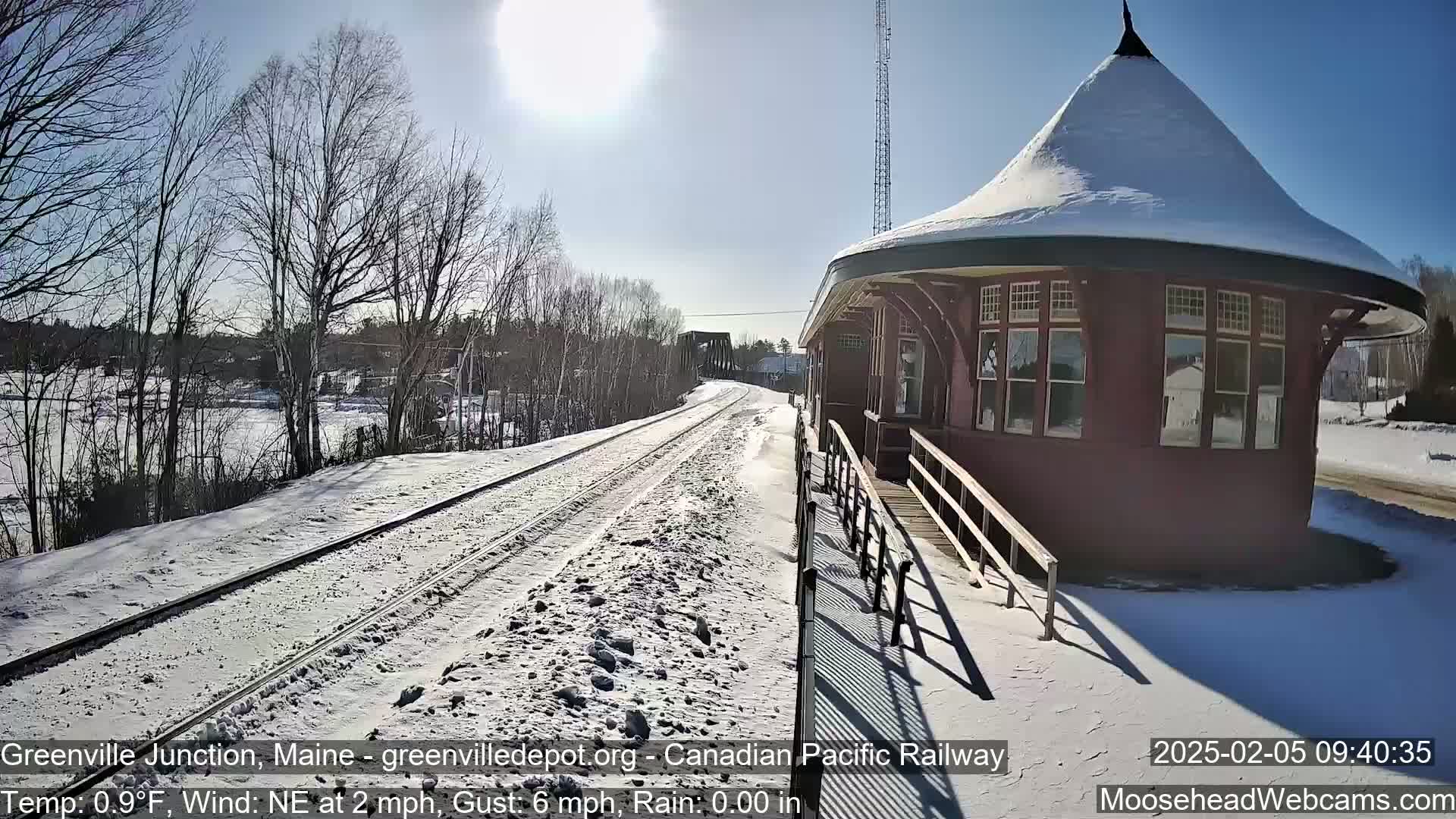 A snow-covered train track leads to a red, snow-capped train station on a sunny day with a light northeast wind.