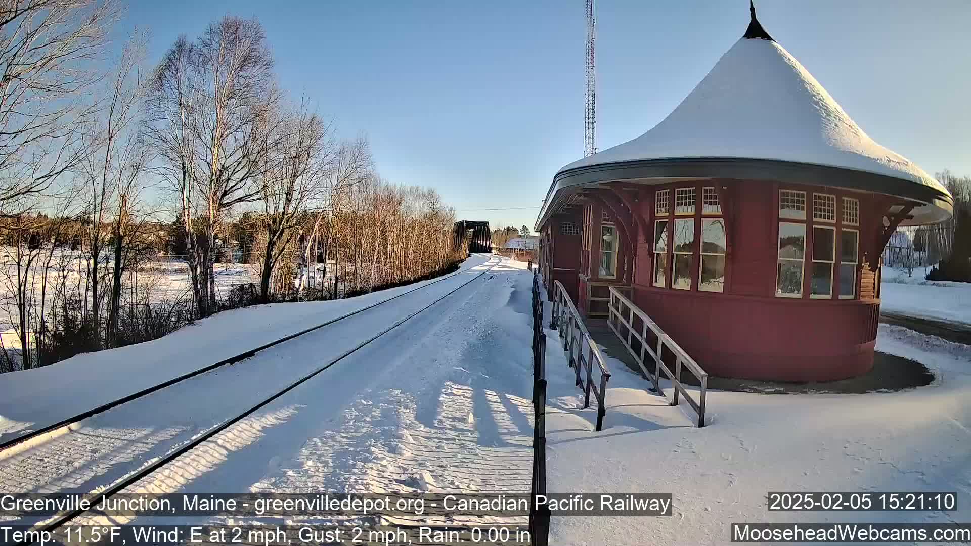 A snow-covered train station with a round, red structure stands beside a snow-covered railway track under a sunny sky.
