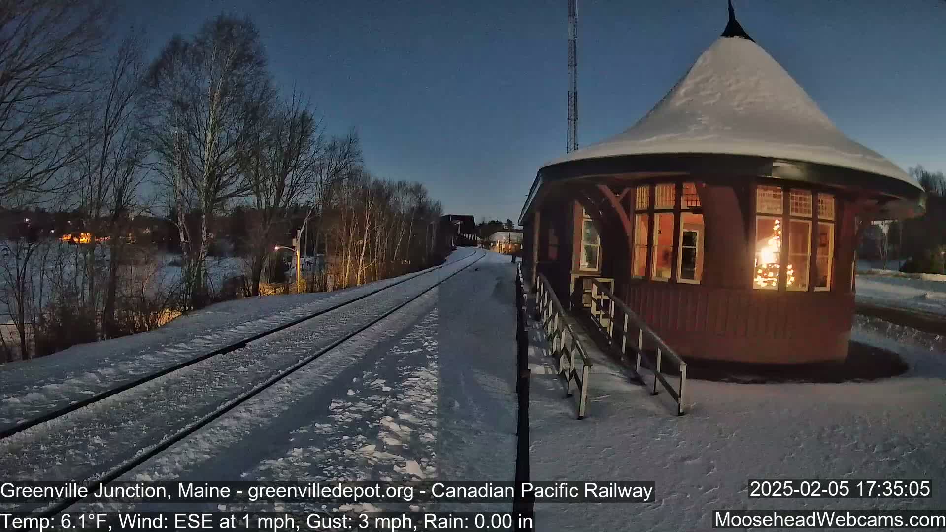 A snow-covered train platform with a lighted building is seen at dusk, with a calm wind.