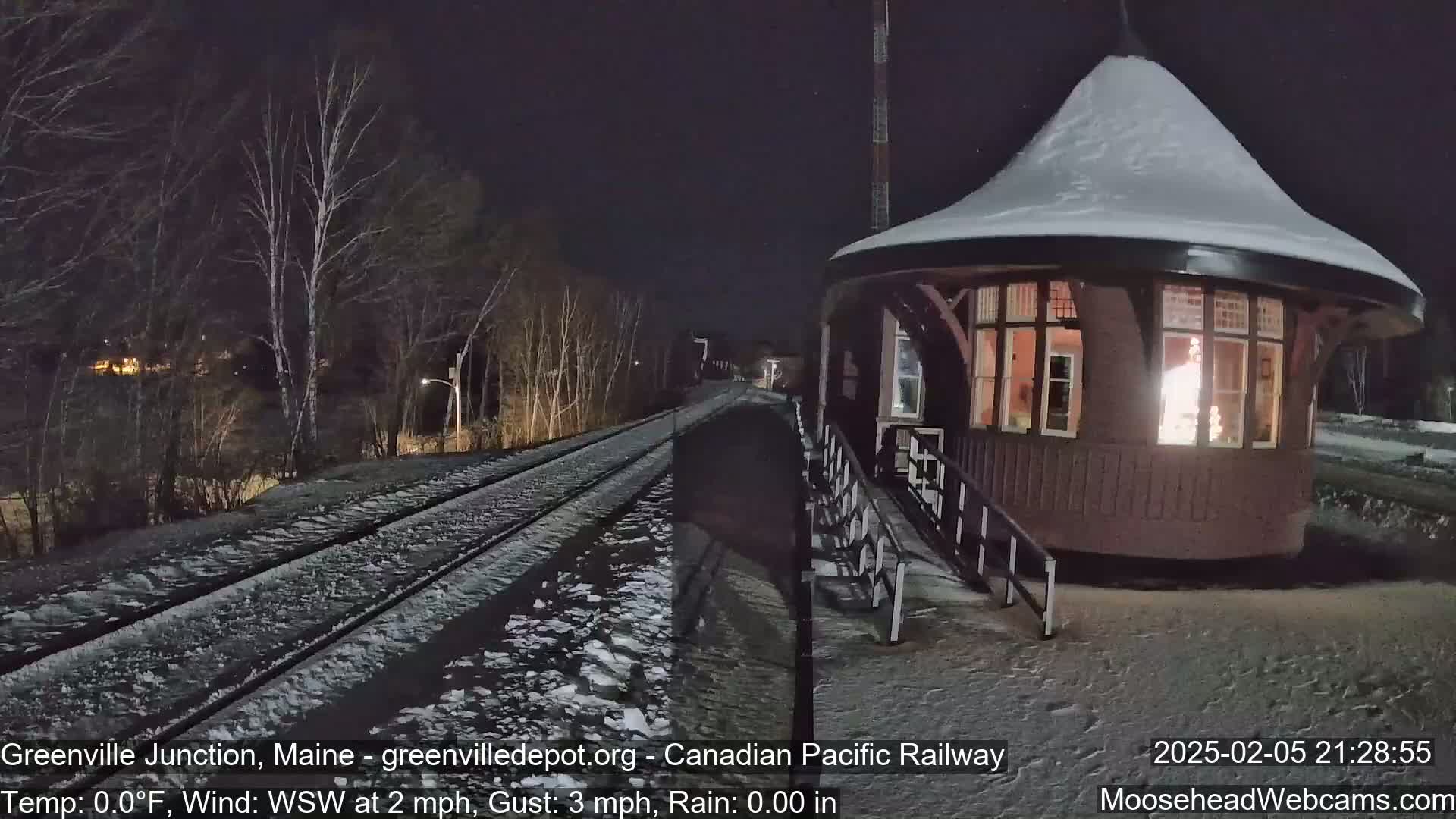 A snow-covered railway track leads to a small, illuminated, snow-covered building at night.