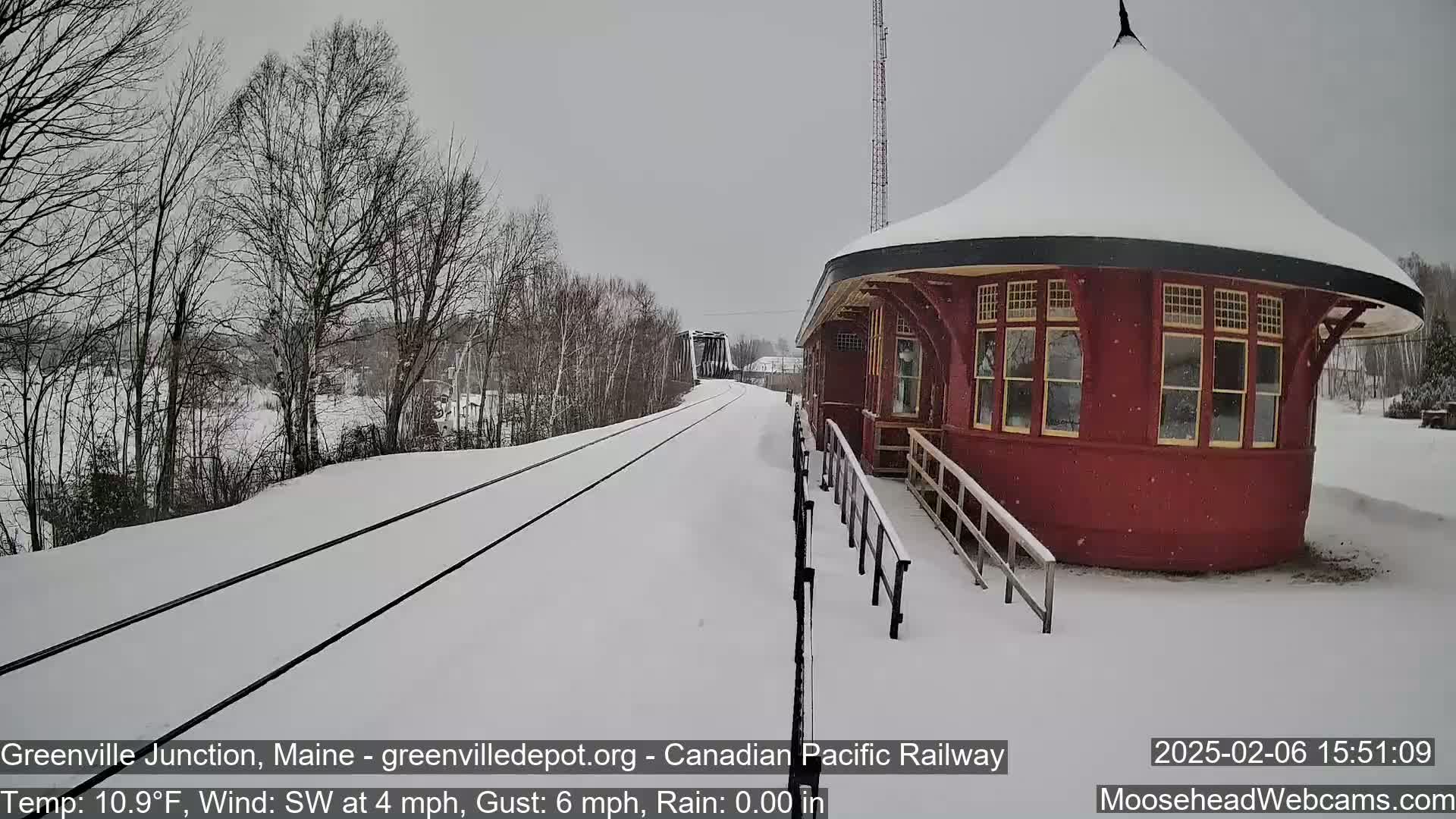 Snow-covered railroad tracks extend toward a red, round train depot under a gray sky, with bare trees lining the tracks.