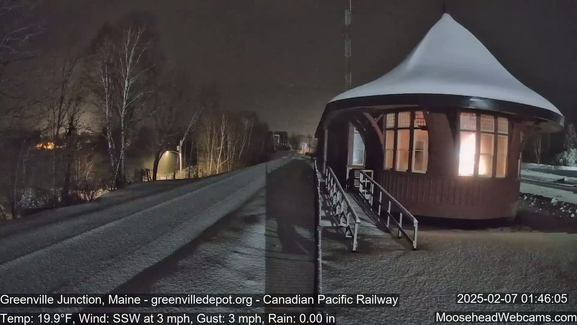 A snow-covered road leads to a small, illuminated, snow-covered building at night under a dark sky with a light wind.