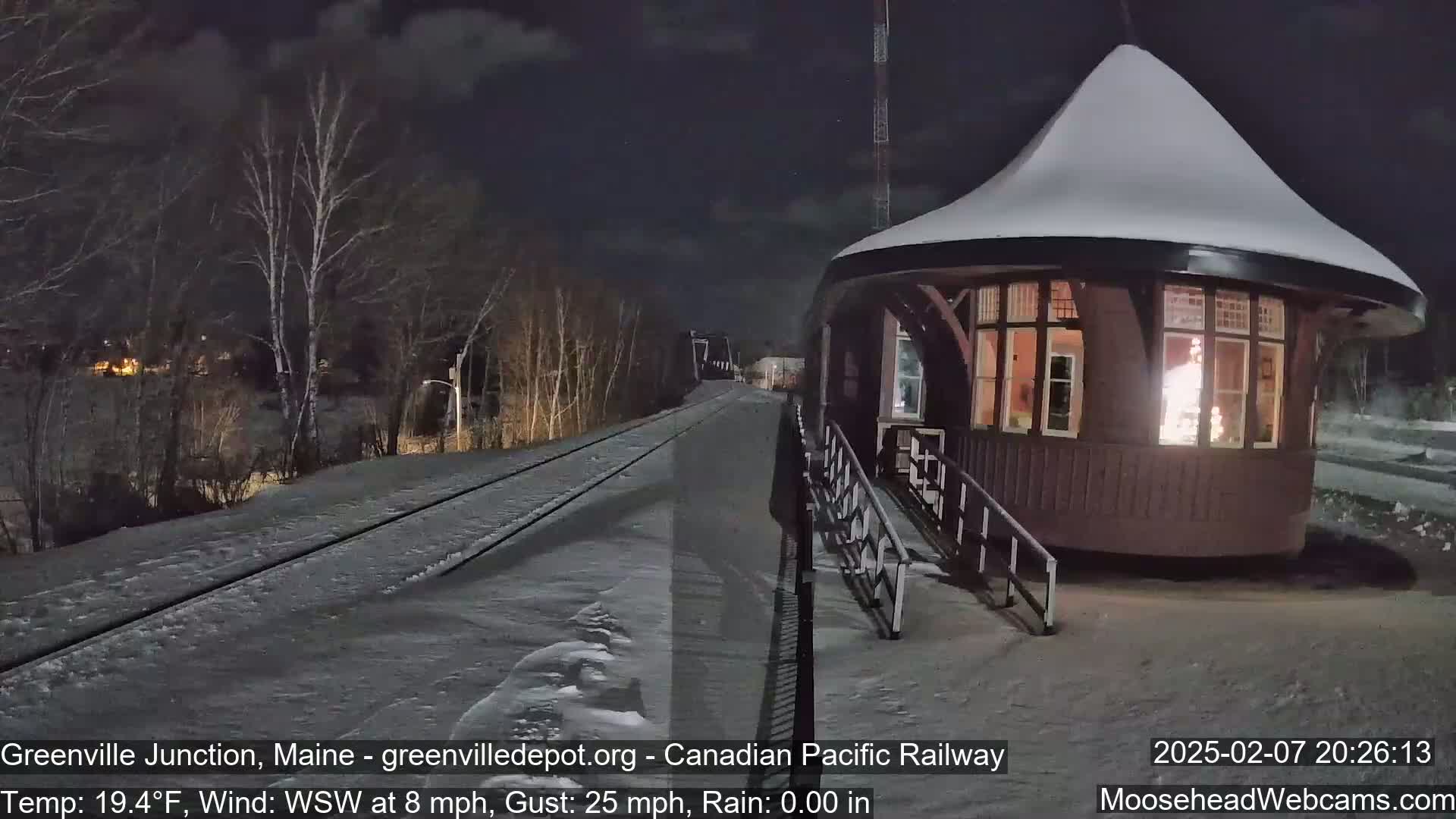A snow-covered train depot is illuminated at night, with a snow-covered railway extending into the distance under a dark, windy sky.