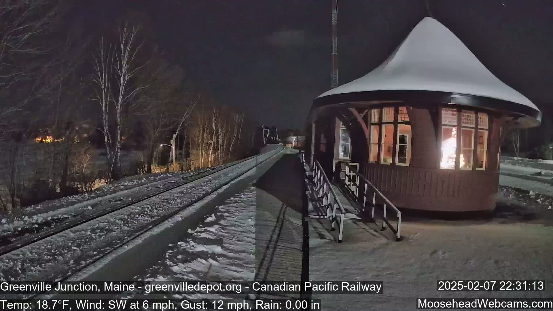 A snow-covered train station with a lit interior sits beside a snow-dusted railway track at night.
