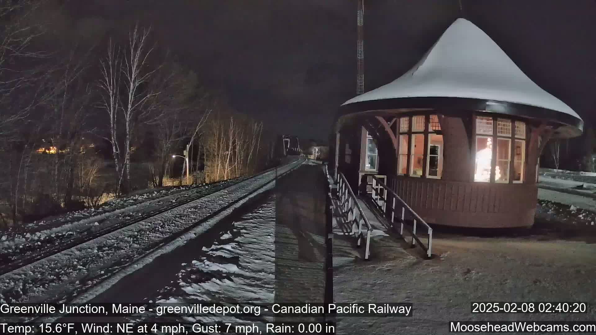 A snow-covered train depot is illuminated from within, situated beside a snow-dusted railway track at night, under a northeast wind.