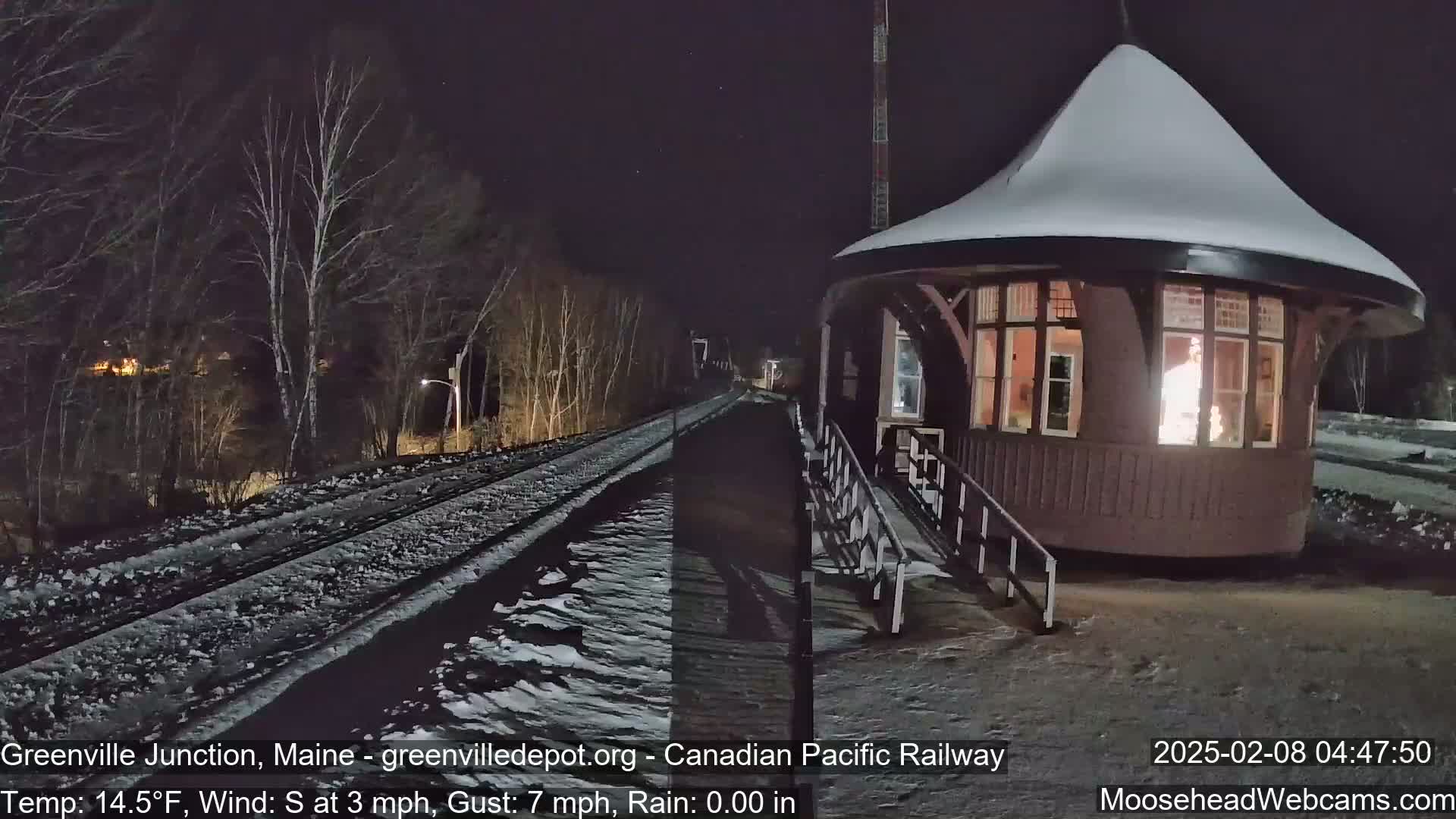 A snow-covered train platform with a lit-up building is seen at night, with a light dusting of snow on the tracks and surrounding ground.