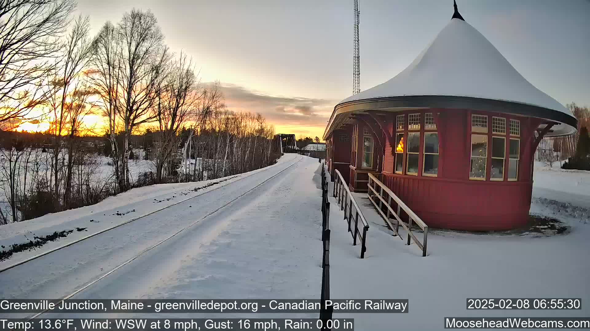 A snow-covered train platform and red circular building are situated beside a snow-covered railway track during a sunset with a light wind.