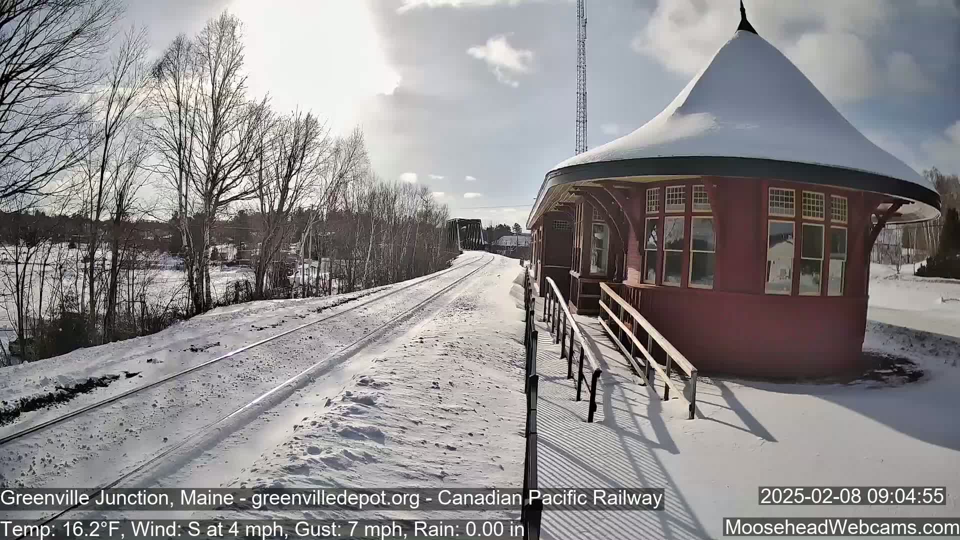 A snow-covered train platform with a red, round depot building is visible under a partly sunny sky.