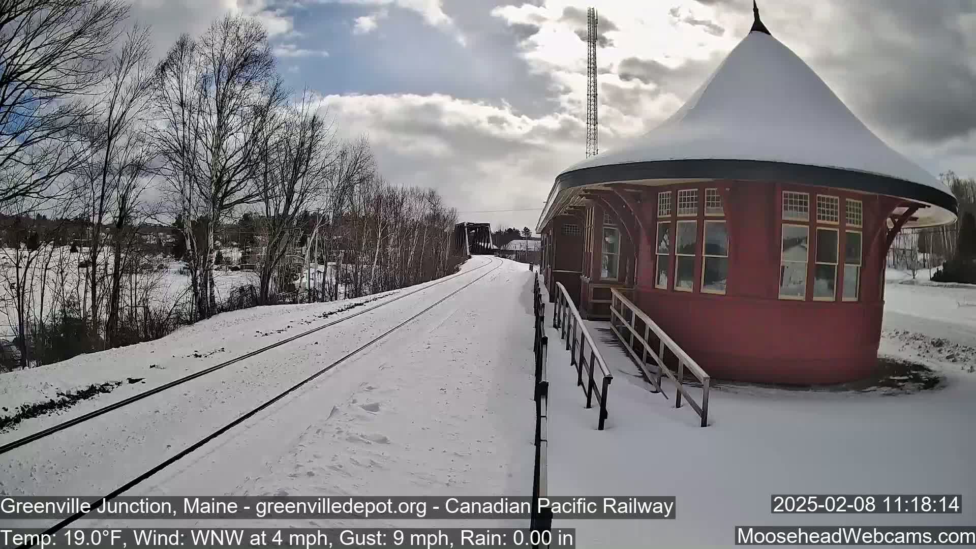 A snow-covered train platform with a red, round train station building is visible, with train tracks extending into the distance under a partly cloudy sky.