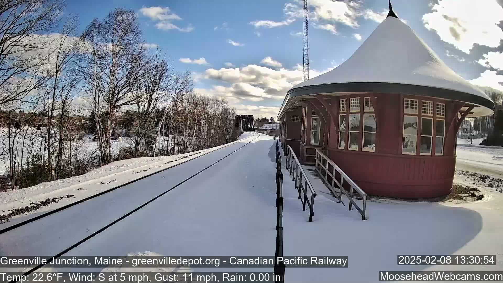A snow-covered railroad track leads to a red, round train station under a partly sunny sky.