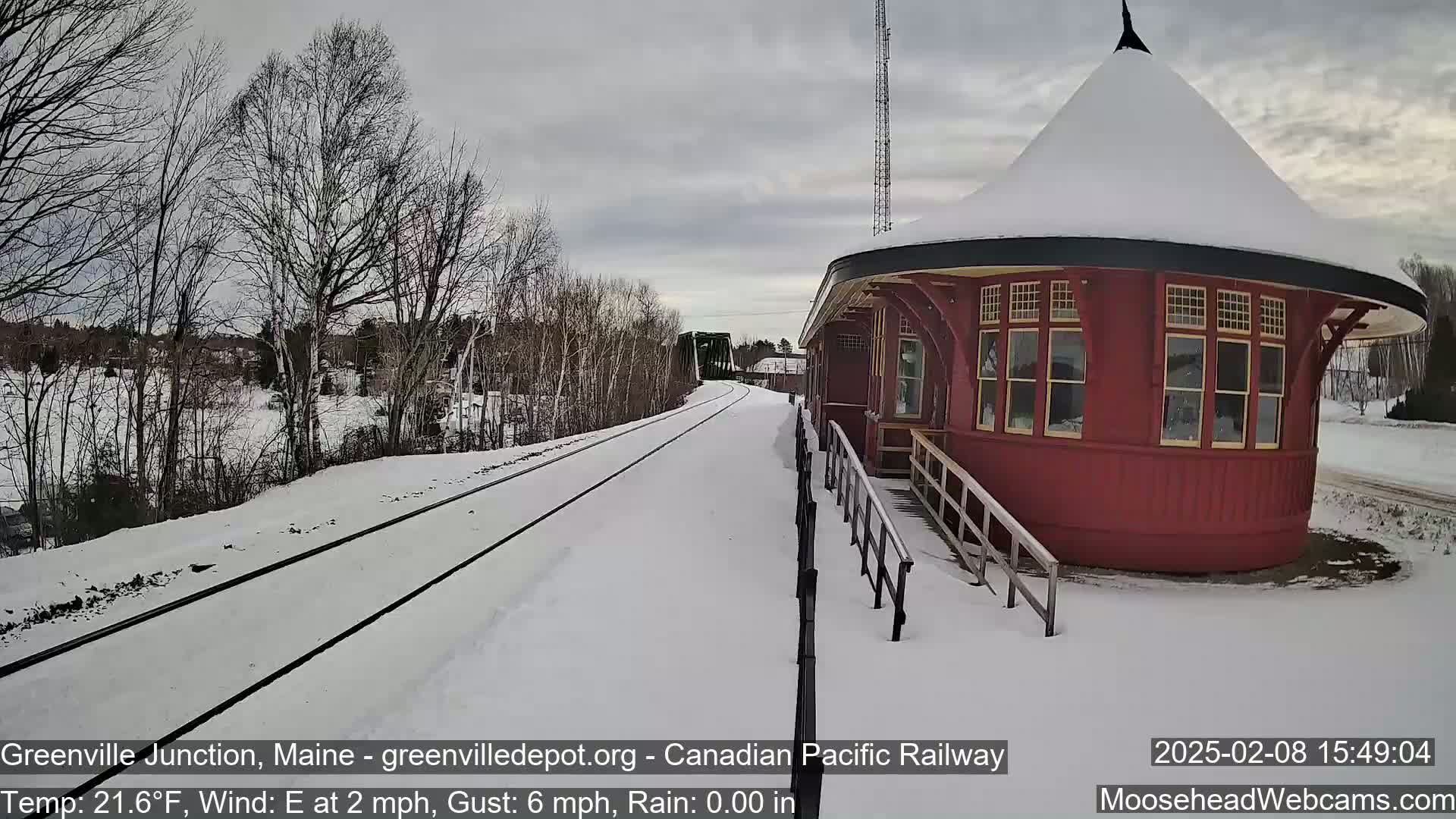 A snow-covered train station with a round red building and tracks extending into the distance under a cloudy sky.