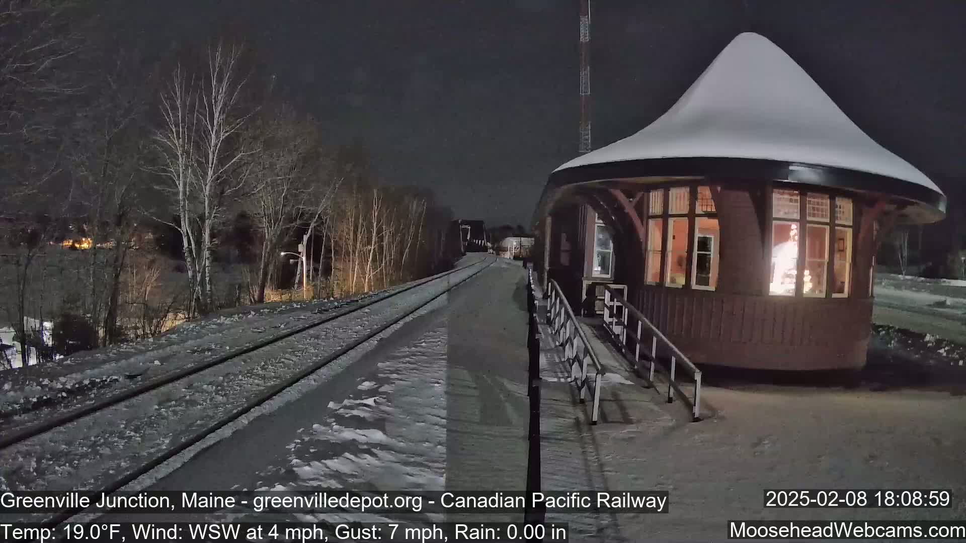 A snow-covered train platform with a small, snow-covered building next to train tracks under a dark night sky.