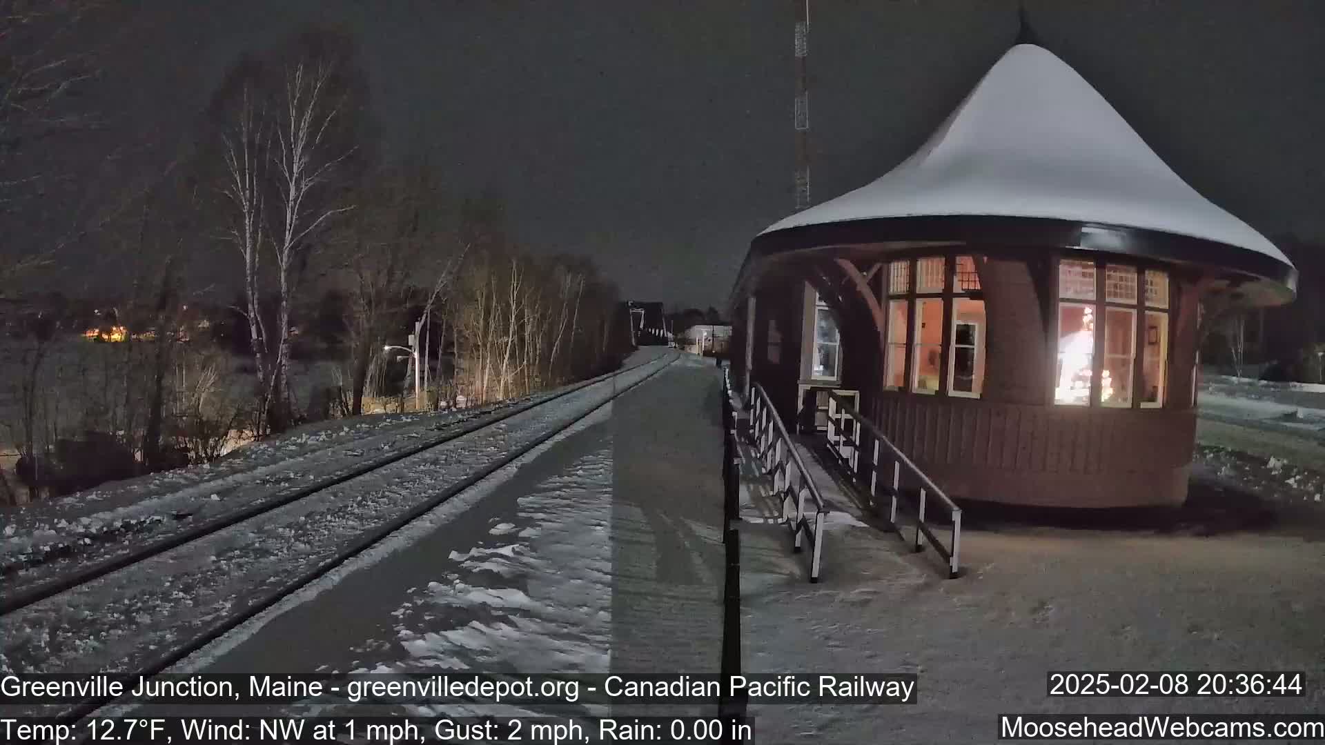 Snow-covered train tracks and a snow-dusted, illuminated train station are visible at night under a dark sky.