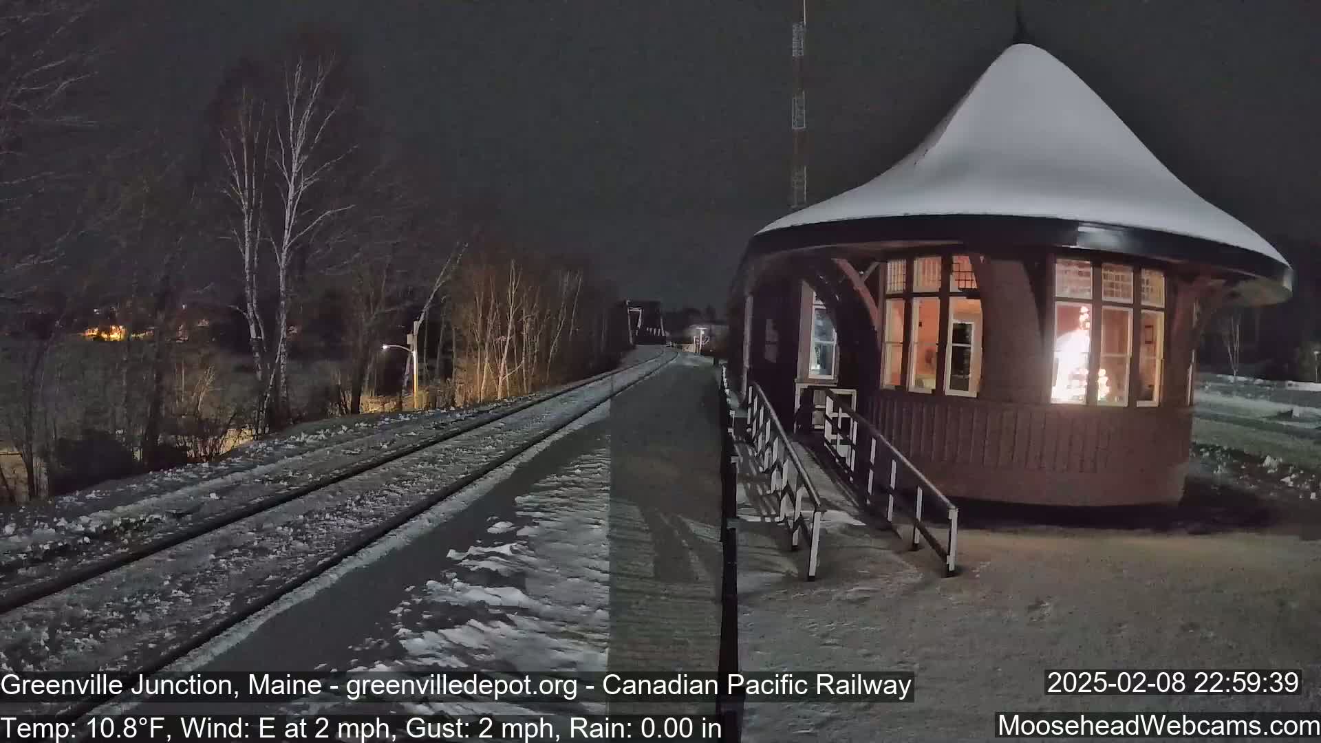A snow-covered train station with a lit interior is situated beside a snow-dusted railway track at night under a dark sky.
