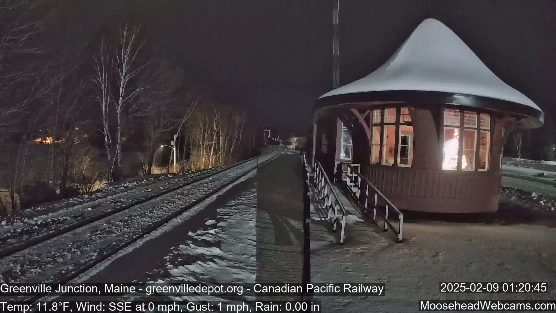 A snow-covered, round train depot with illuminated windows sits beside a snow-dusted train track at night under a dark sky.