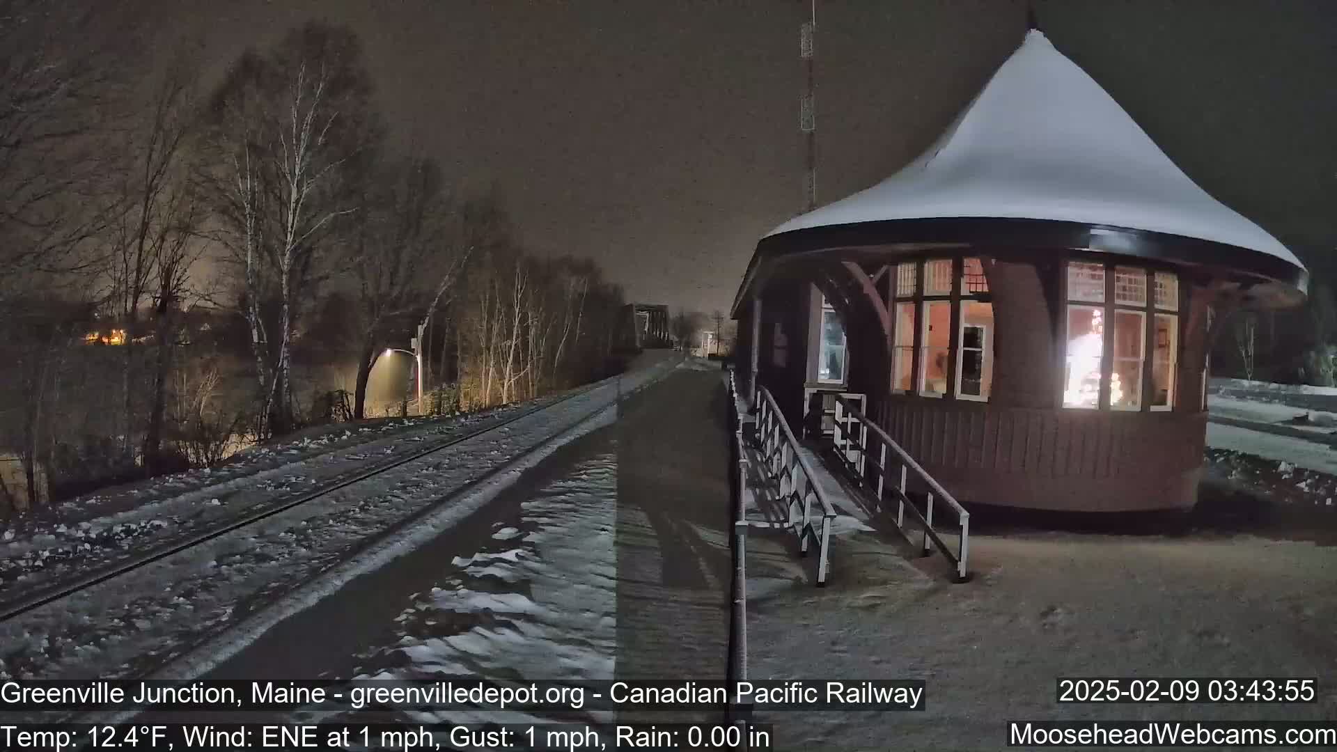 A snow-covered, octagonal train depot is illuminated at night next to a snow-dusted train track under a dark sky.