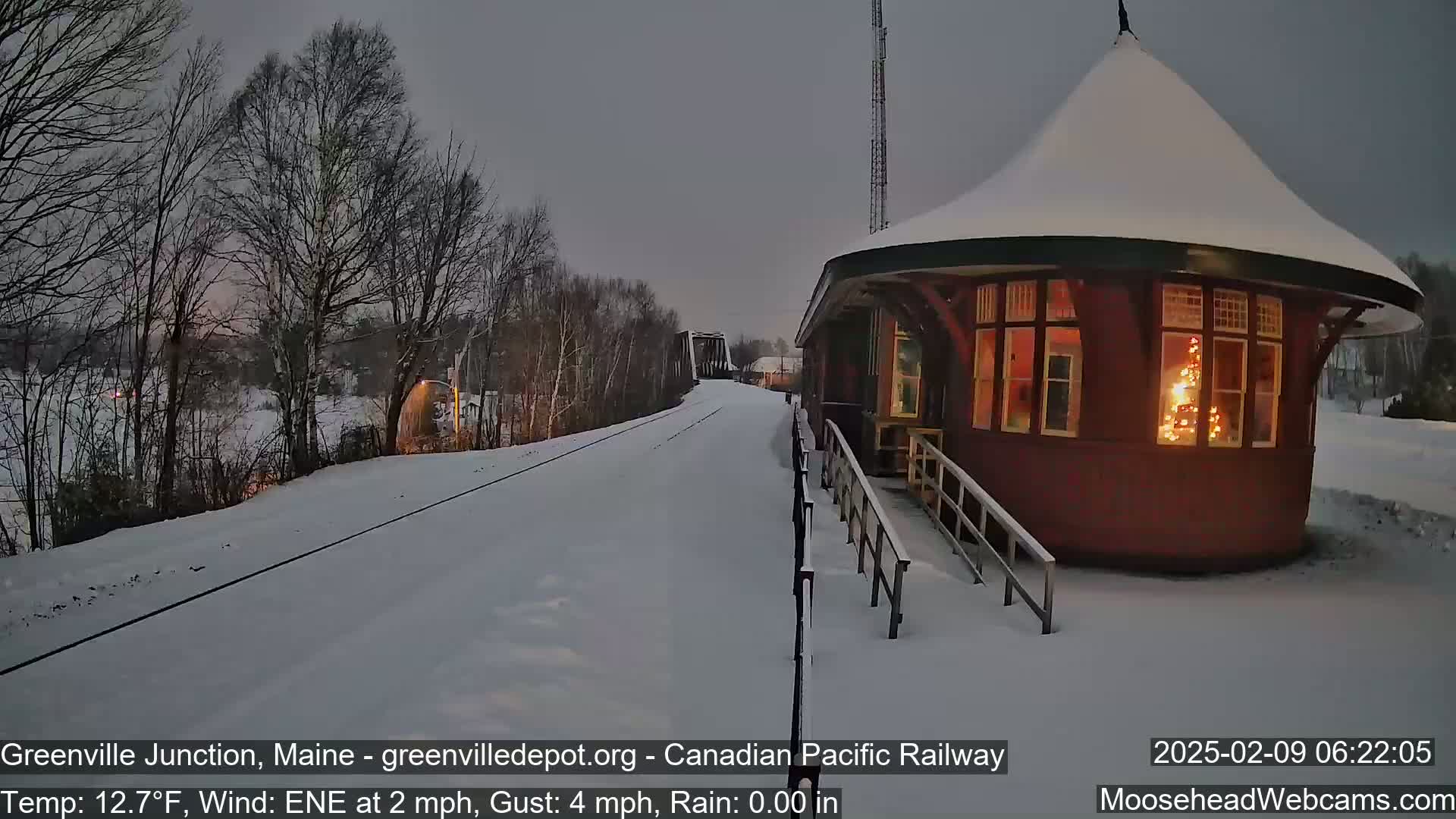 A snow-covered train platform with a small, illuminated building next to railway tracks under a twilight sky.