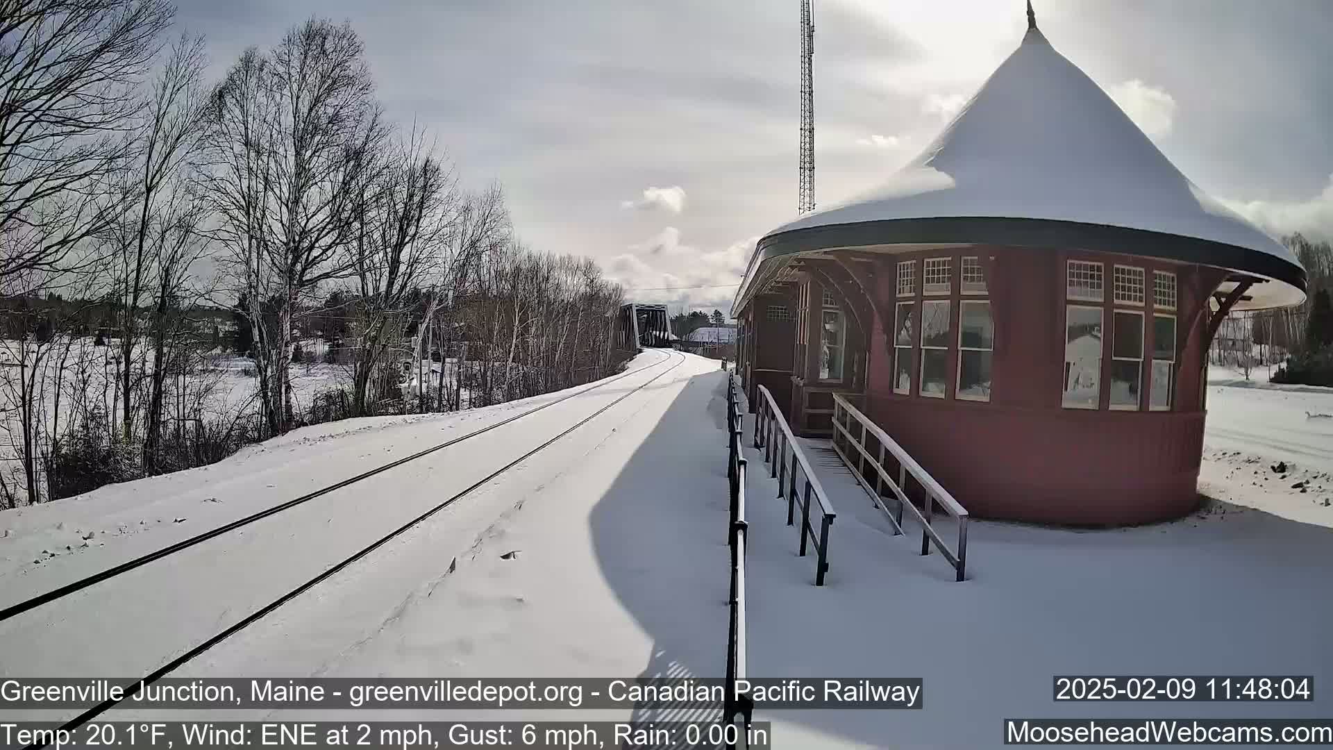 A snow-covered train station and railway tracks are visible on a sunny, cold day.