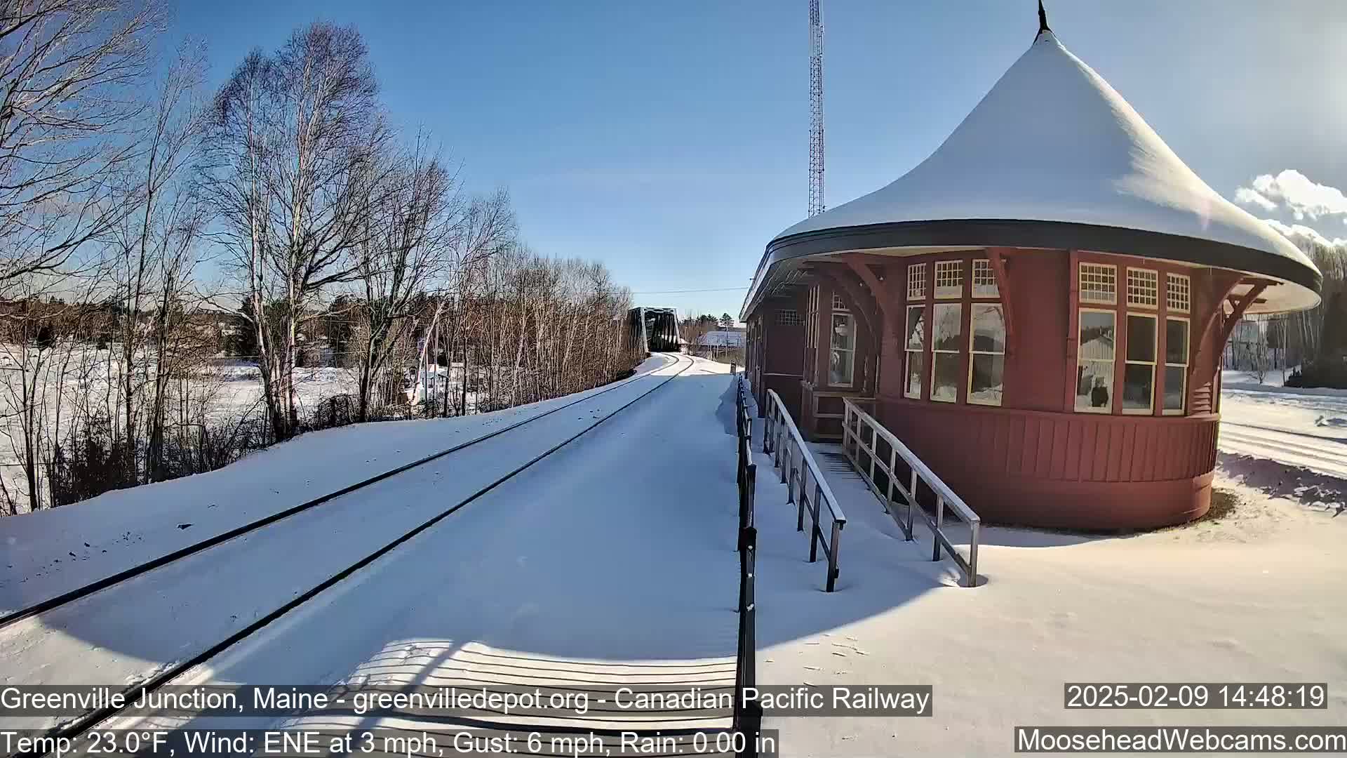 Snow-covered train tracks extend toward a red, snow-dusted train station under a clear blue sky.
