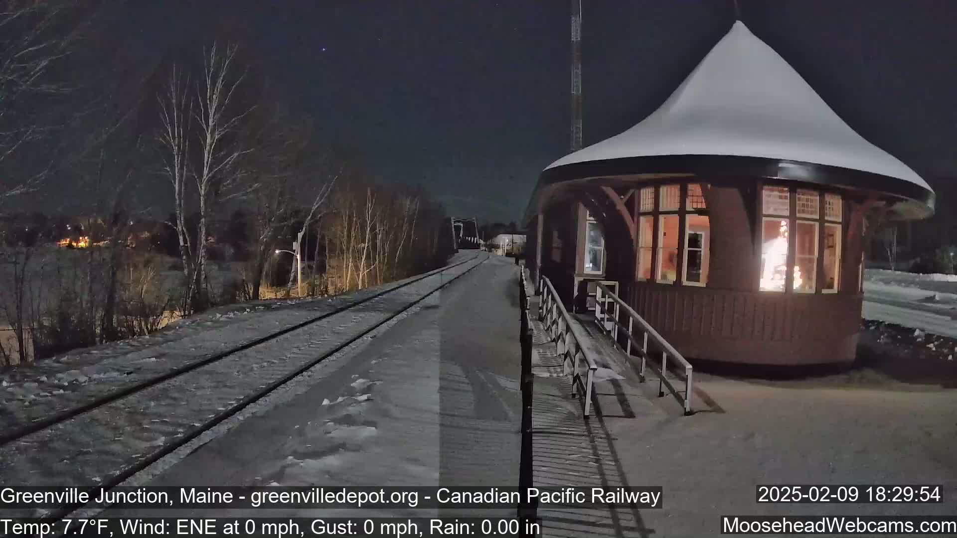 A snow-covered, illuminated train depot stands beside a snow-covered railway track under a dark, clear night sky.