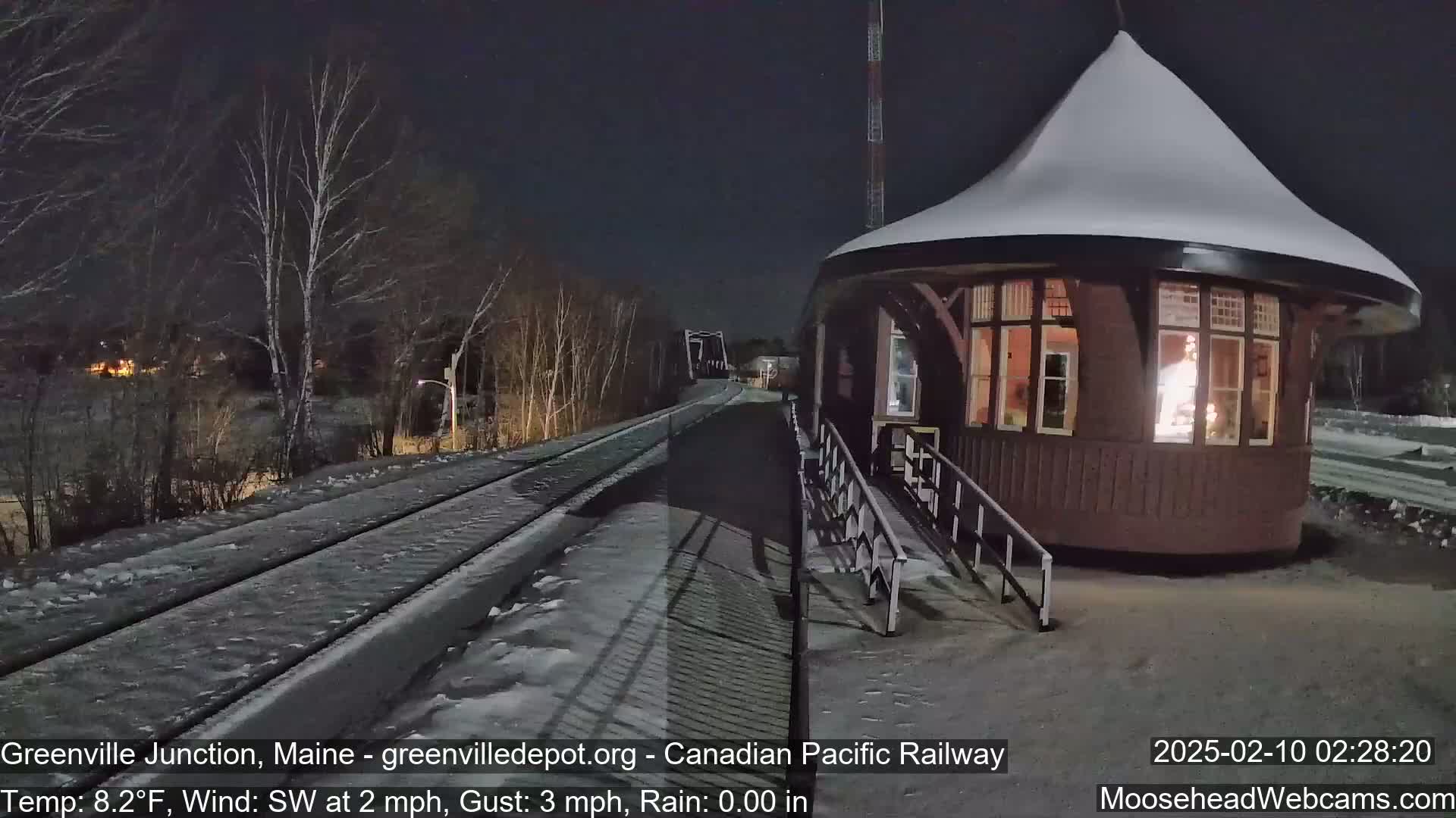 A snow-covered train station with lights on inside sits next to a snow-covered railway track under a dark night sky.