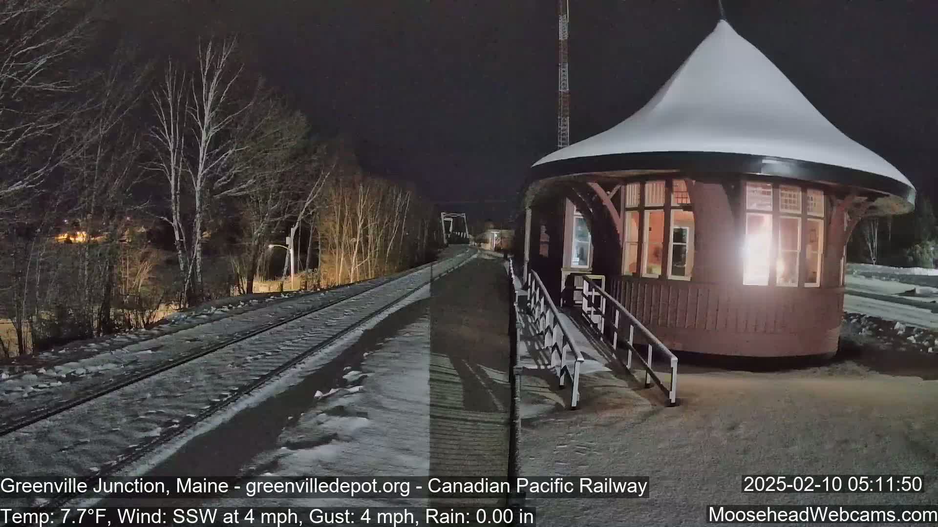 A snow-covered train station and railway tracks are illuminated at night under a dark sky, with a light wind.