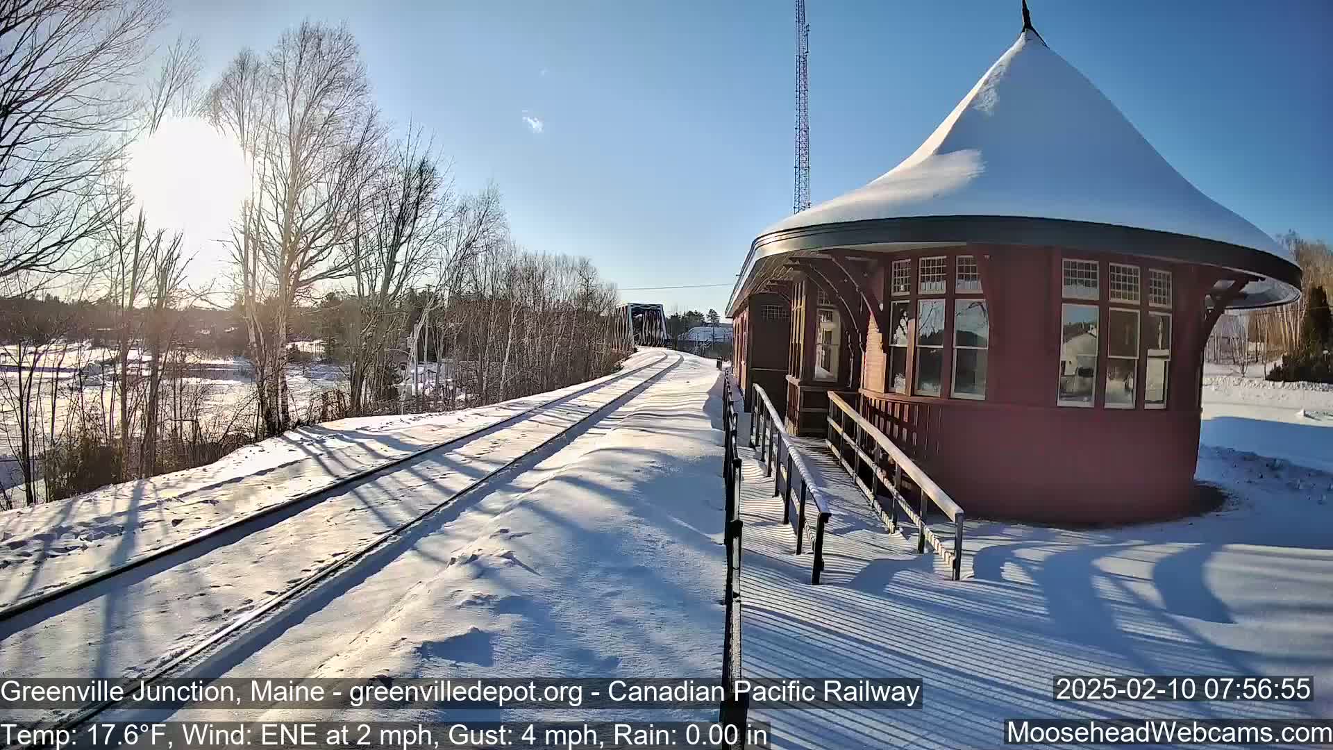 A snow-covered train station with a circular roof stands next to snow-covered train tracks on a sunny day with a light wind.