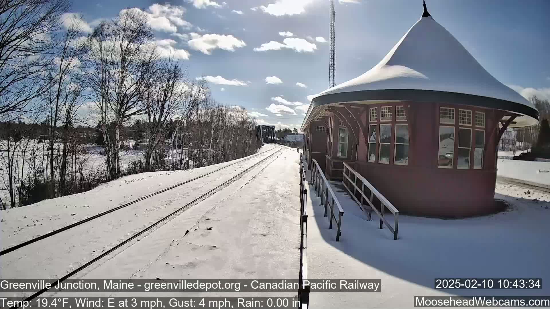 A snow-covered train platform with a red, octagonal building and train tracks extending into a sunny winter landscape.