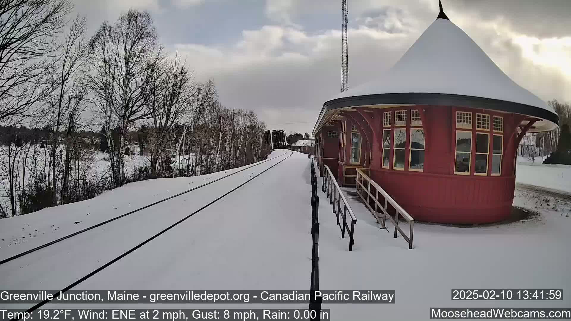 Snow-covered railroad tracks lead to a red, round train depot under a partly cloudy sky.