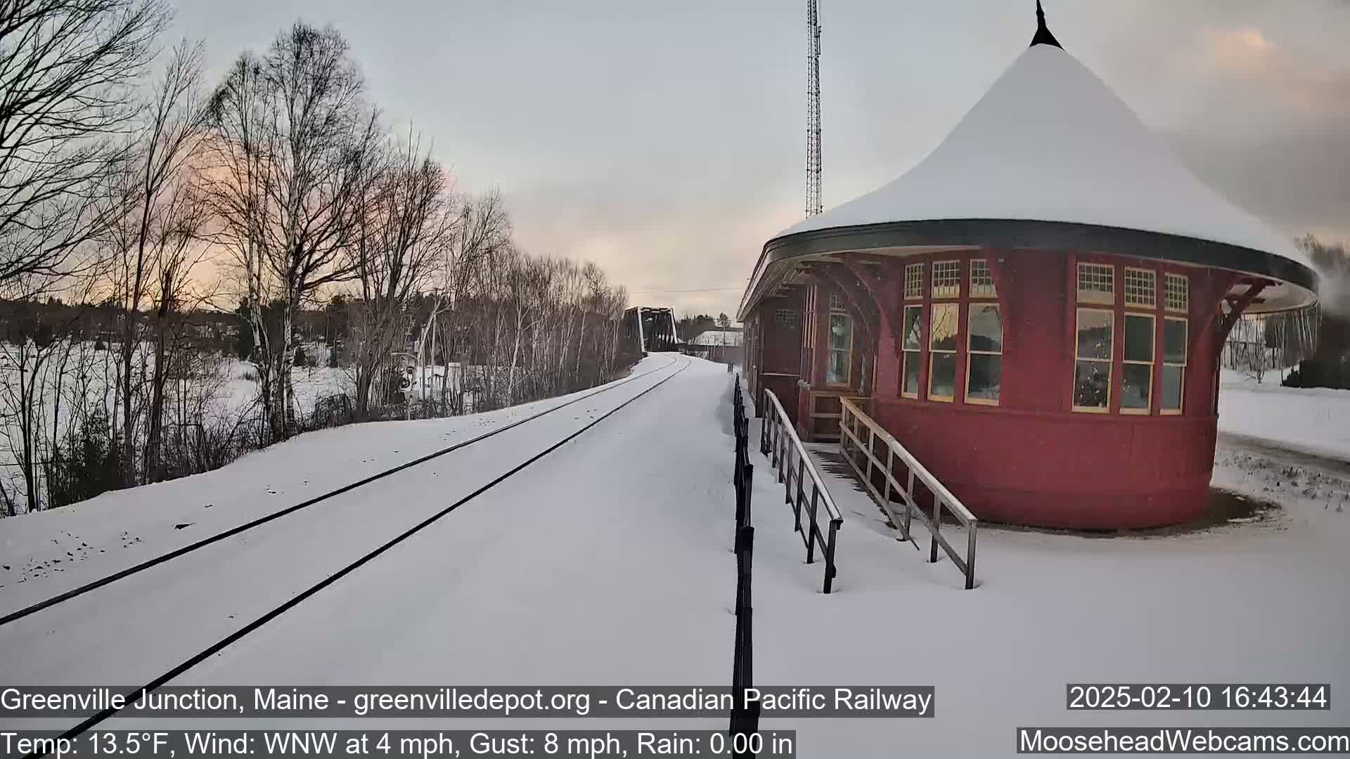 Snow-covered train tracks lead to a red, round train station under a partly cloudy sky.