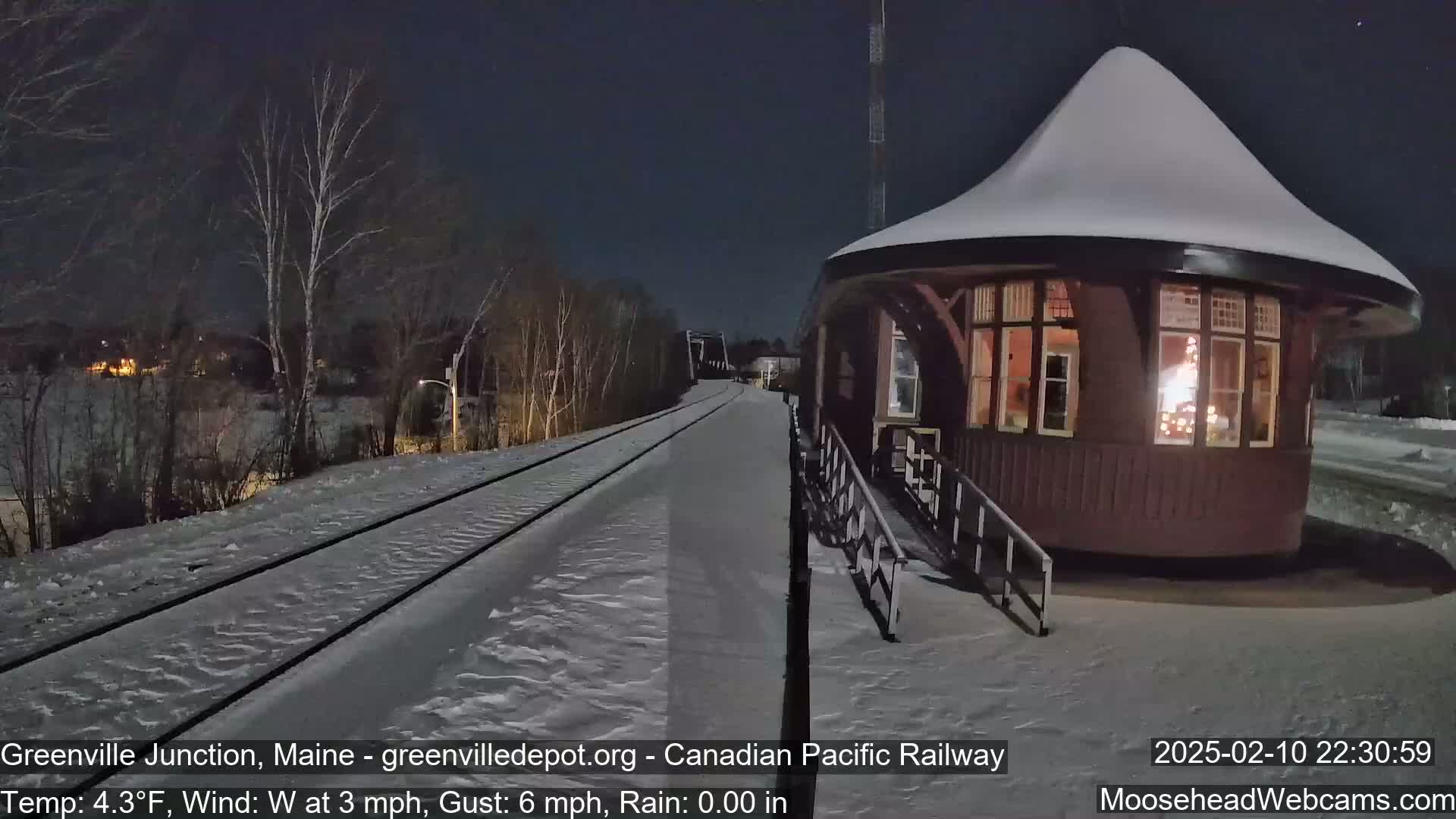 A snow-covered train platform and a round, snow-topped building with illuminated windows are visible at night under a clear sky.
