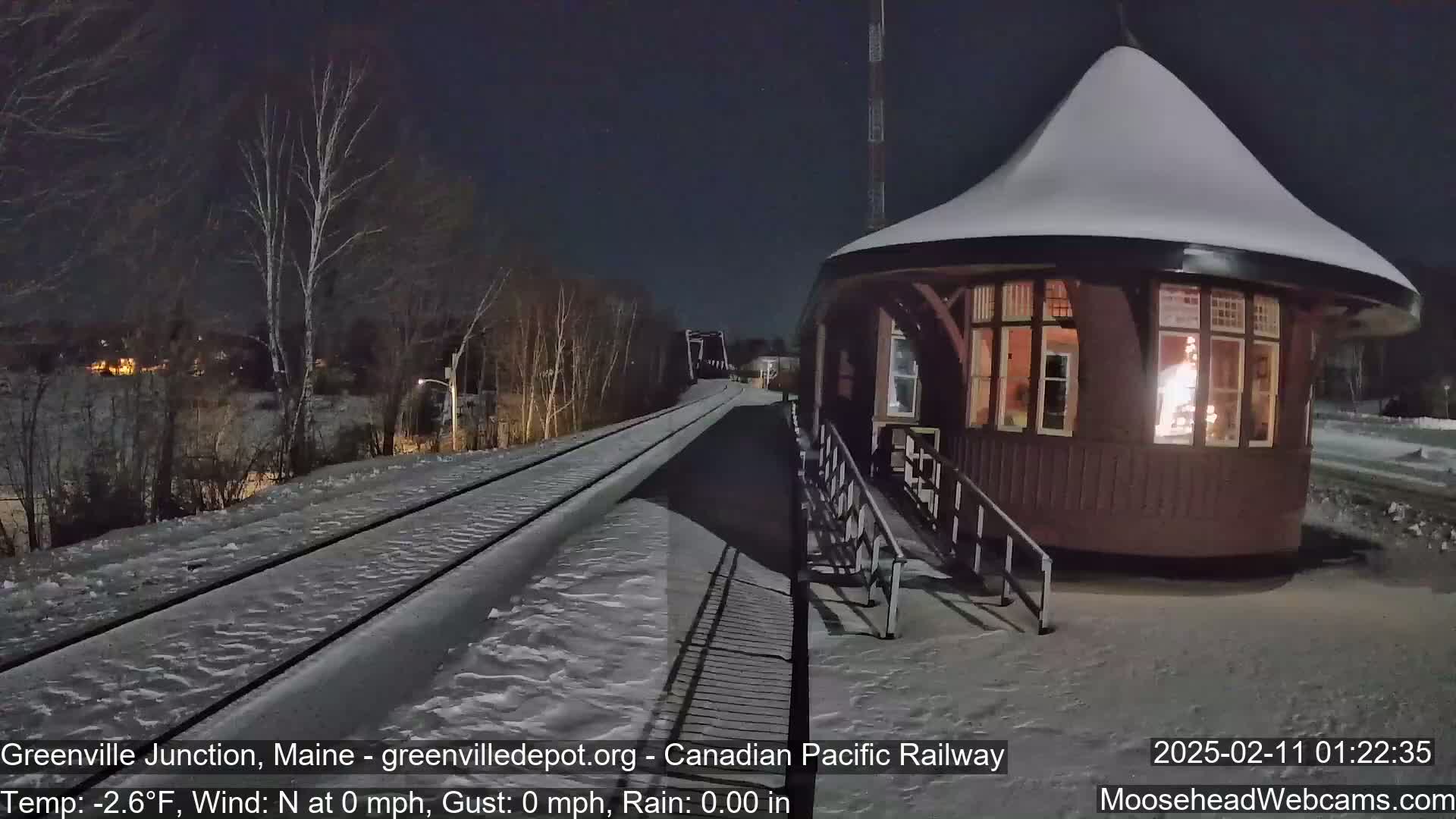 A snow-covered railway platform with a small, snow-capped, octagonal building next to a set of train tracks under a dark, clear night sky.
