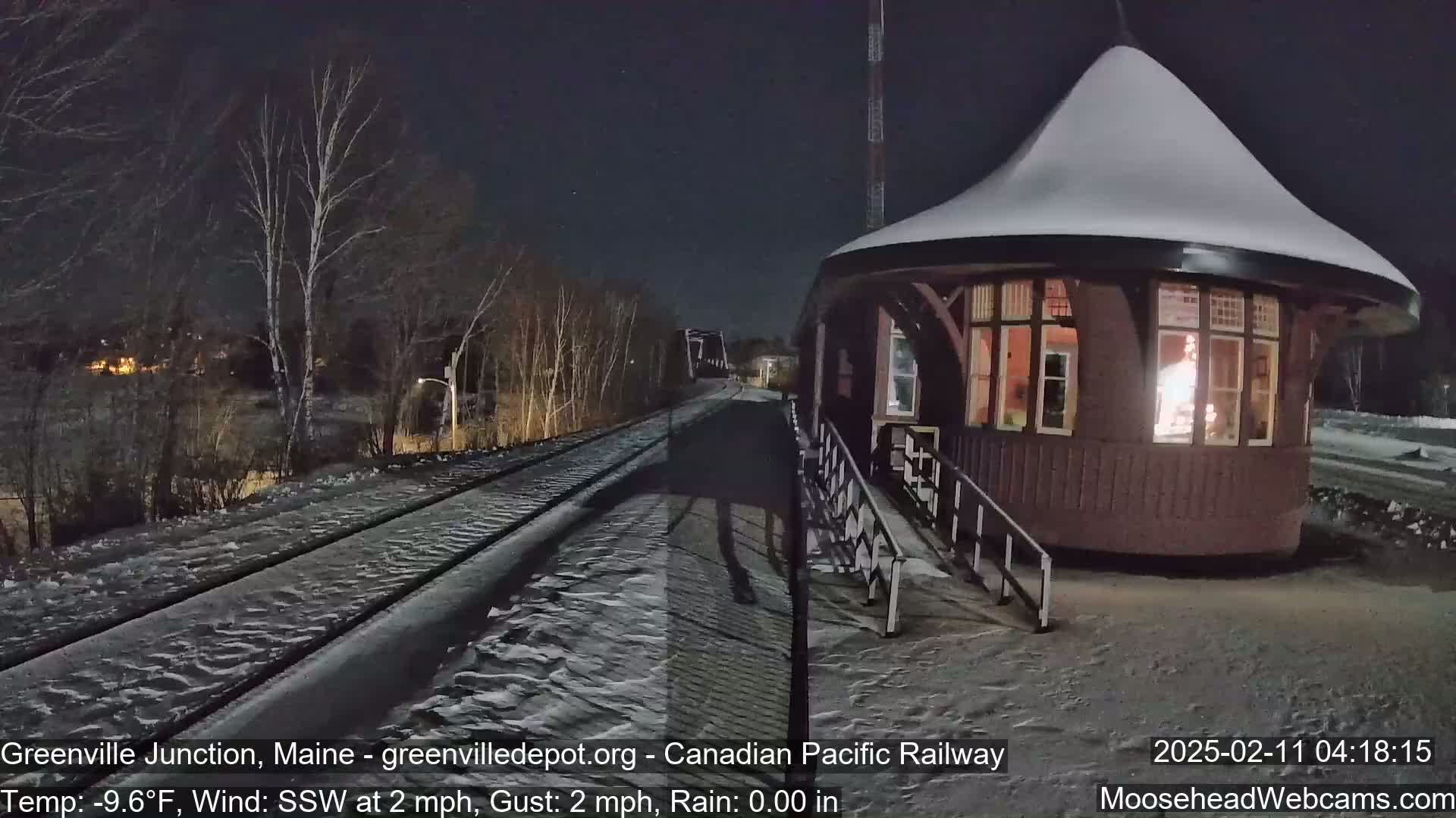 A snow-covered train platform and a small, snow-topped building are illuminated at night next to railroad tracks, under a dark sky with a light south-southwesterly wind.