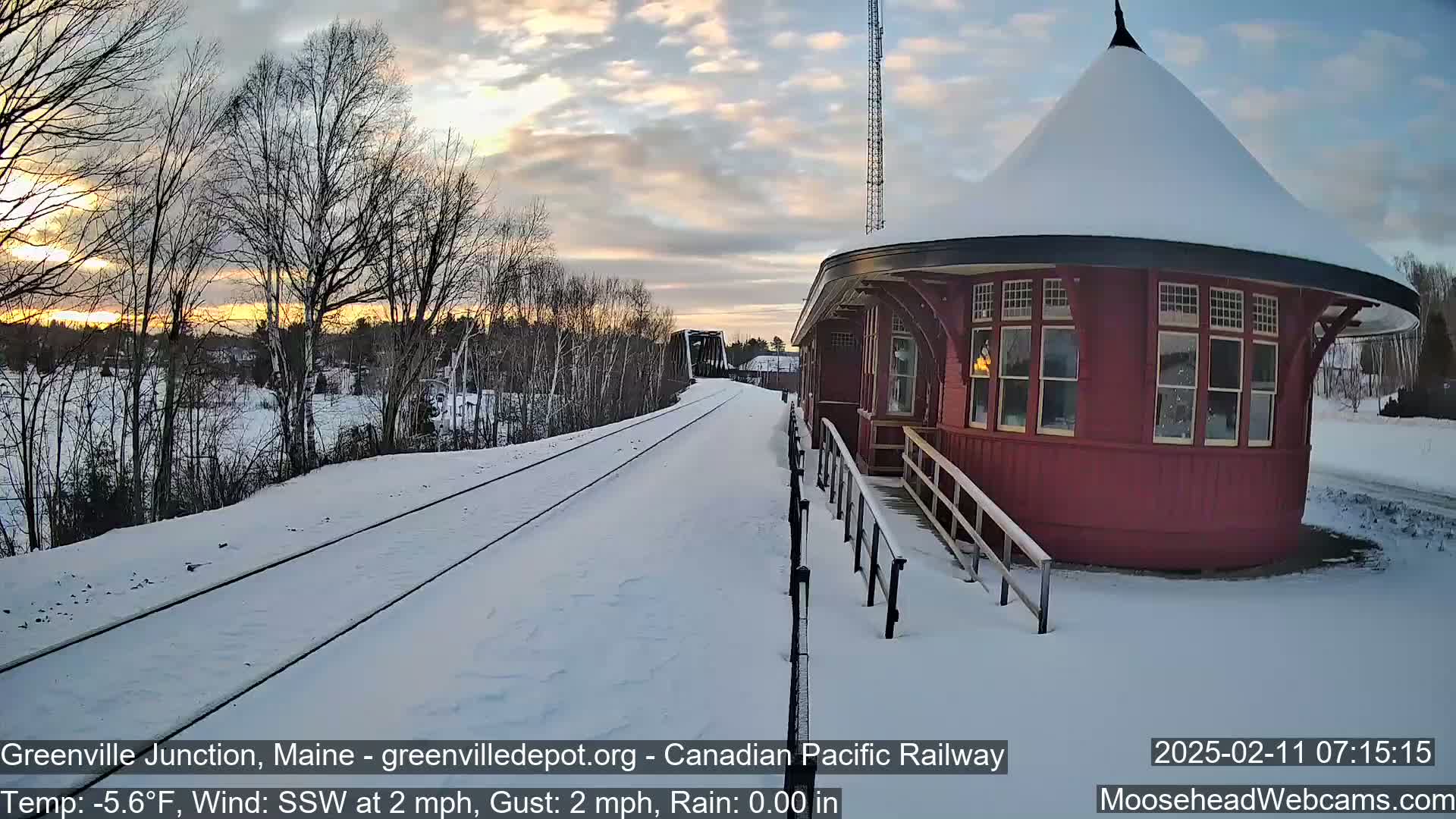 A snow-covered train platform with a red, round train station is visible at sunset, with a train in the distance and snow-covered railroad tracks leading to it.