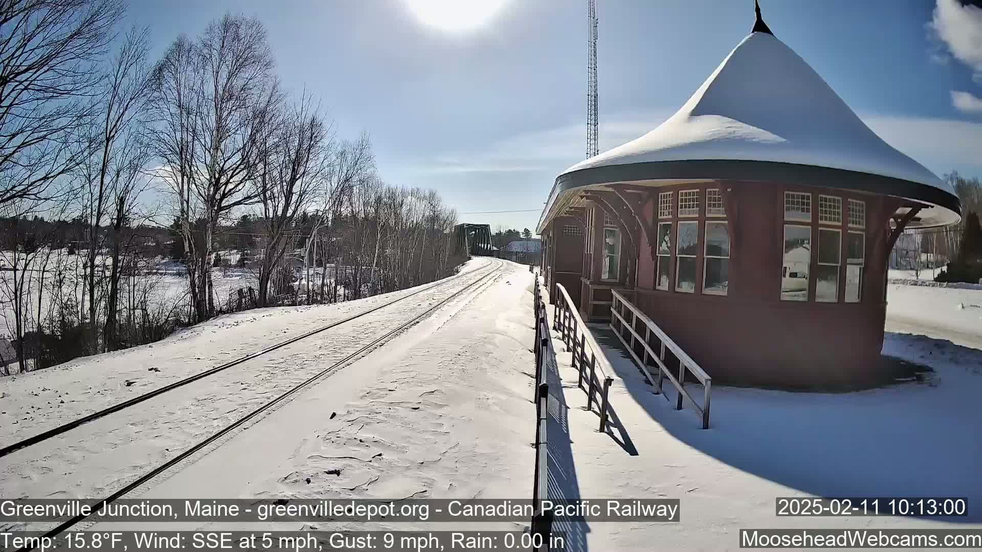 A snow-covered train platform and tracks extend toward a bridge under a sunny sky.