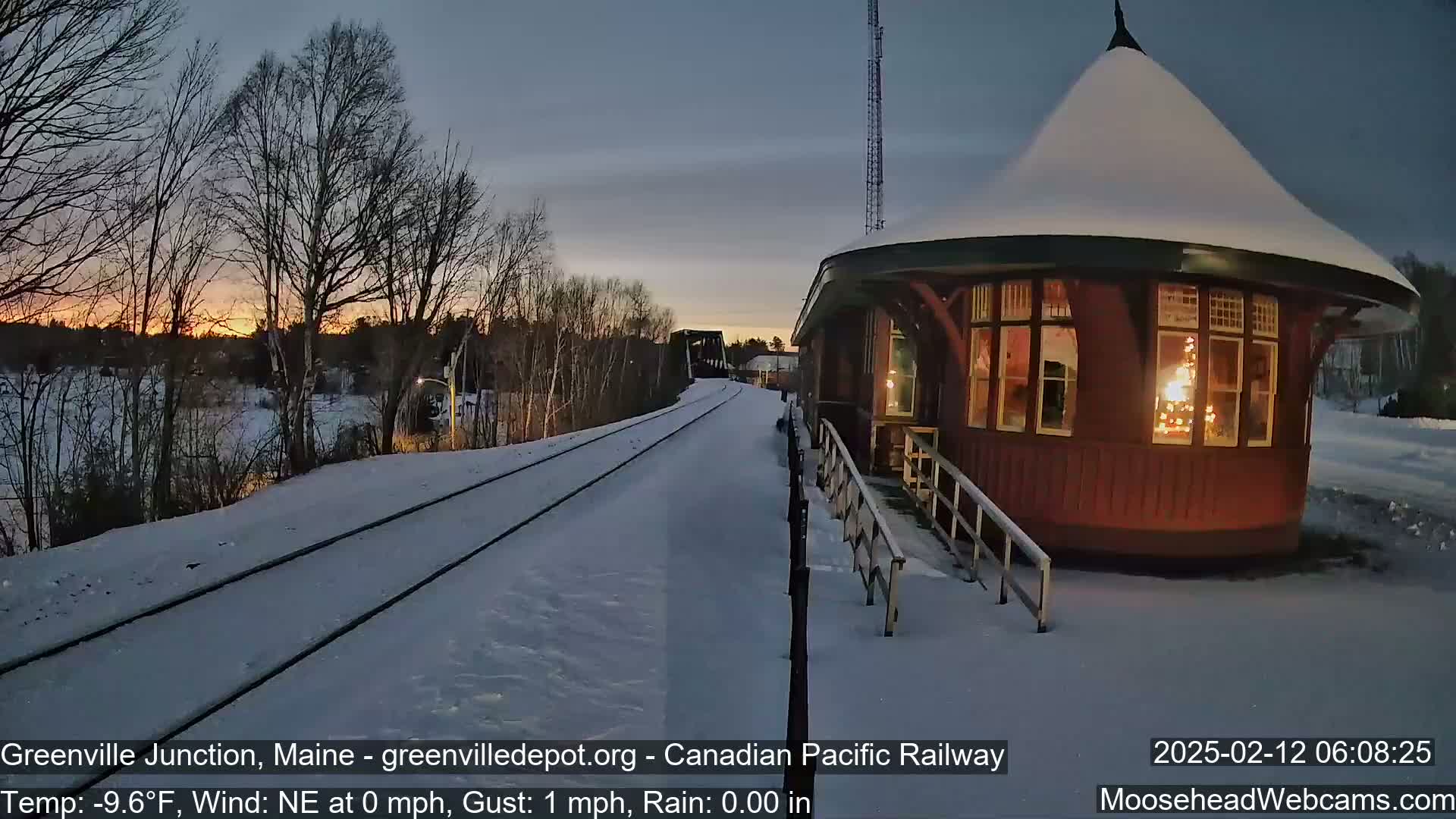 Snow-covered train tracks lead to a snow-dusted, illuminated red railway station during a sunrise with a calm, cold northeastern wind.