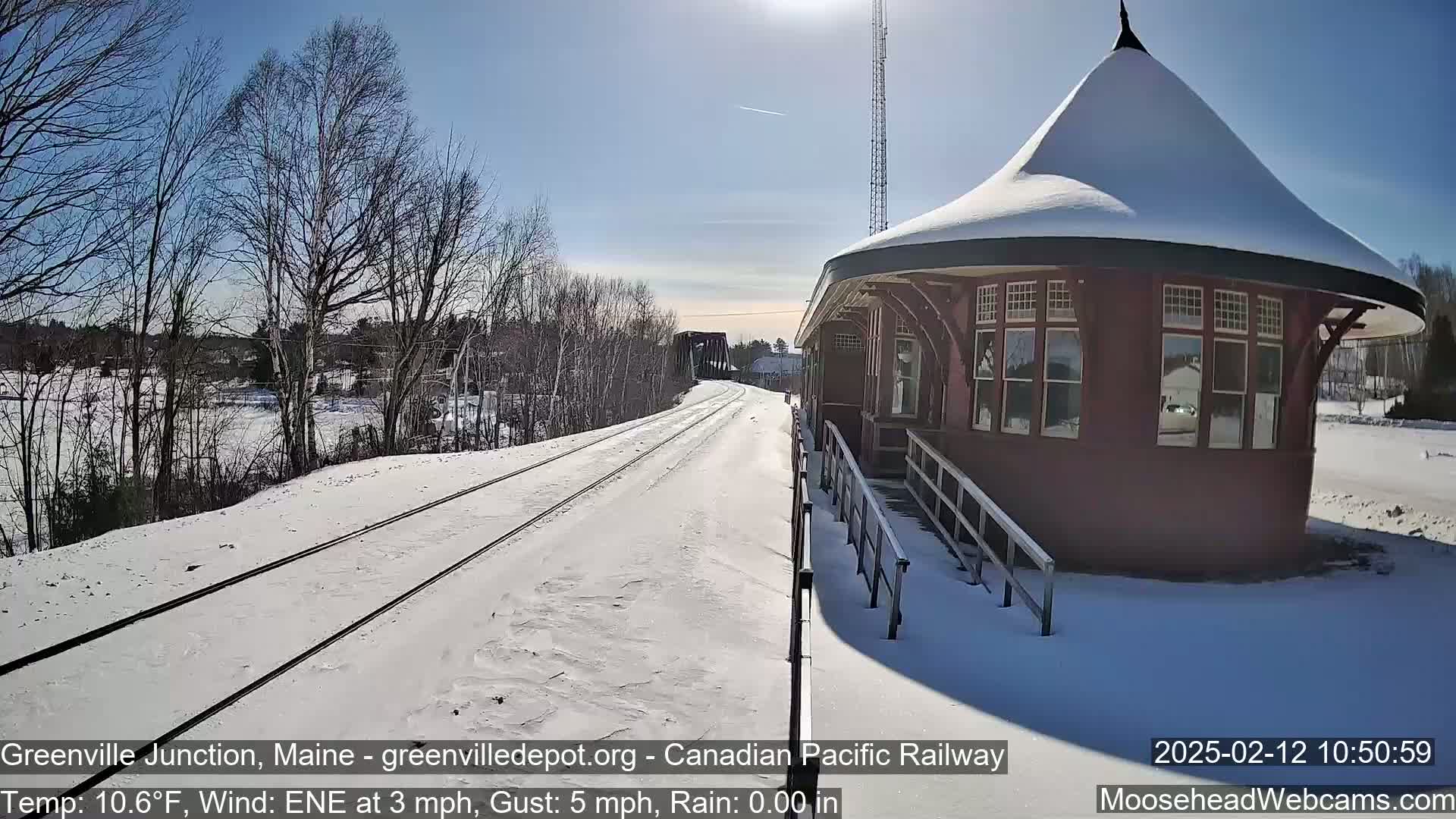 A snow-covered train station platform with railway tracks extending into the distance under a sunny sky.