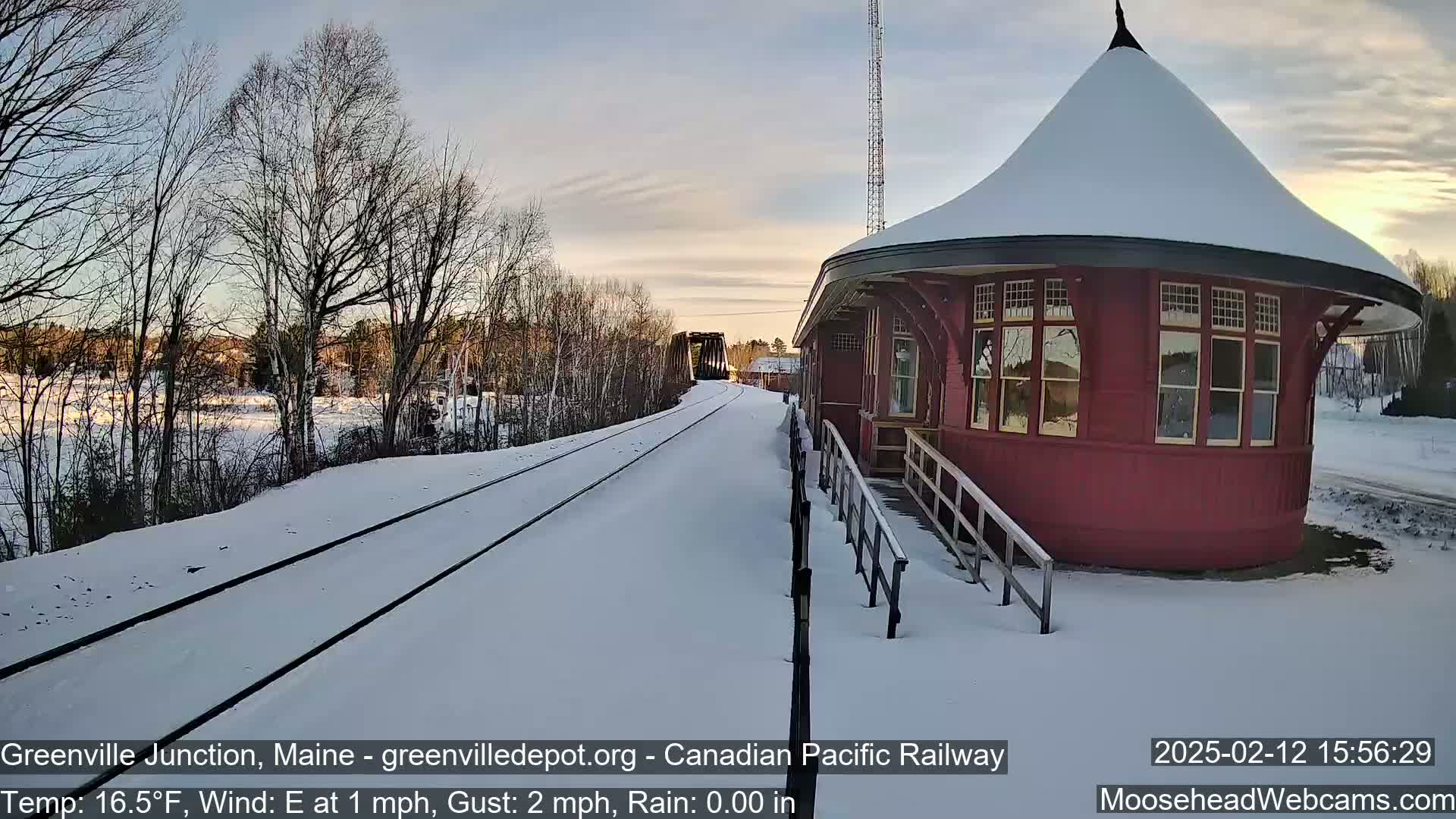 A snow-covered railway track leads to a red, round train depot under a partly sunny sky.