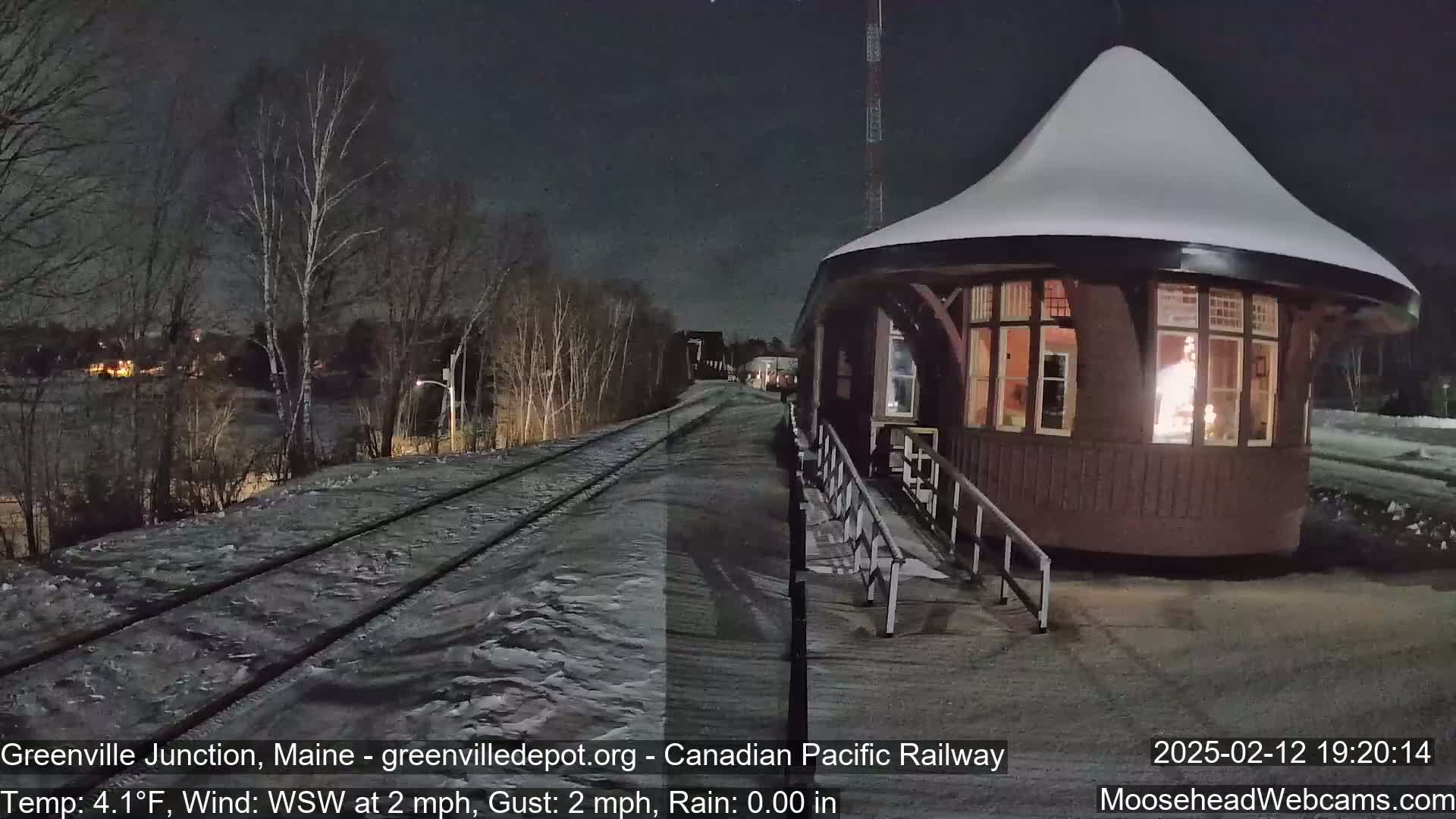 A snow-covered, round train depot with a lit interior sits beside a snow-covered train track at night.