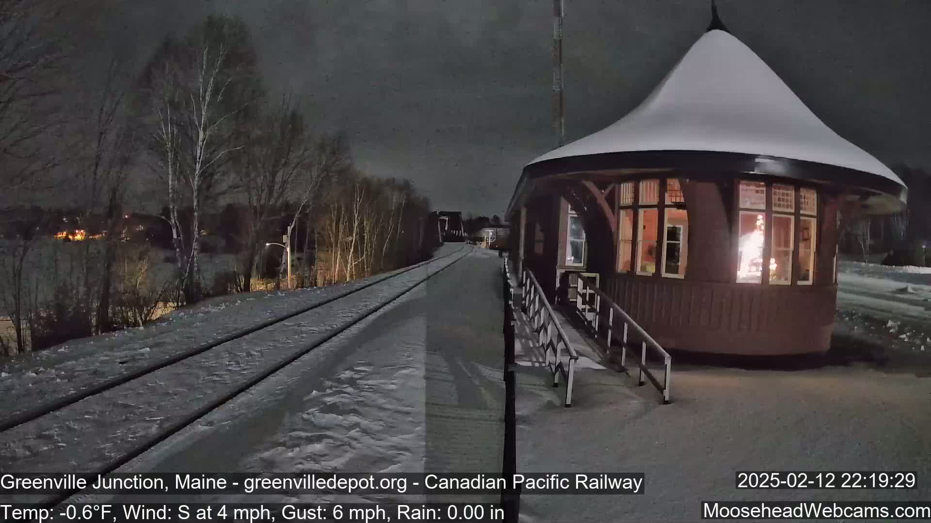 A snow-covered train station platform at night with a dark sky and train tracks extending into the distance.