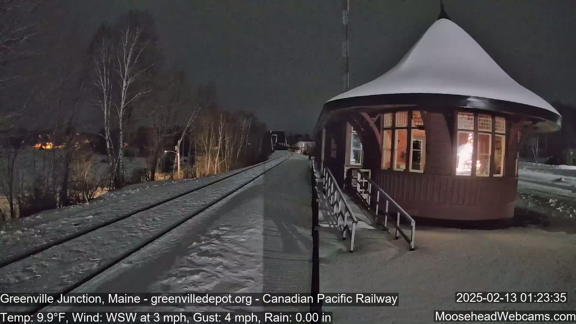 A snow-covered train platform and tracks are illuminated at night, with a small, round, snow-covered building lit from within.