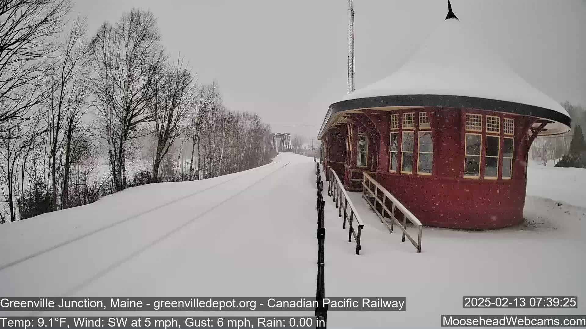 A snow-covered railroad platform and tracks extend toward a bridge under a light snowfall.