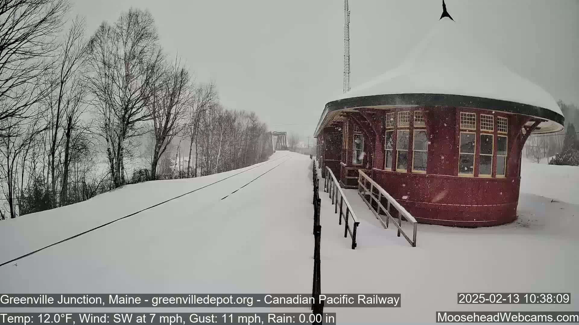 A snow-covered train platform and tracks are visible, with a round, red train station building in the foreground, during a snowfall.