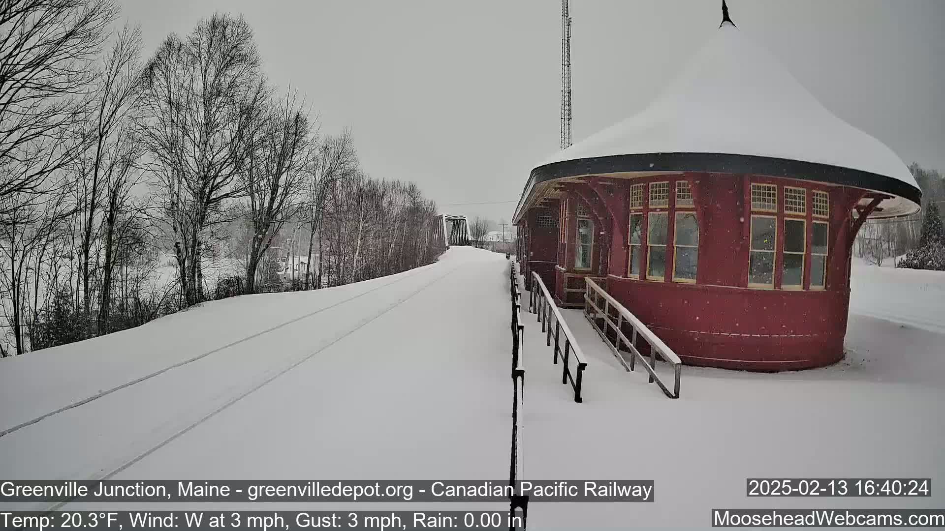 A snow-covered train platform with a red circular building extends from a snow-covered railway track towards a bridge under a lightly snowing gray sky.
