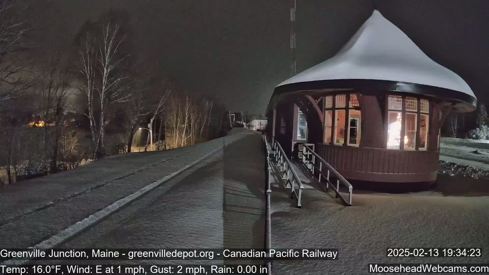 A snow-covered, round train station with a lit interior is seen at night, with a snow-covered road extending into the background under a dark sky.