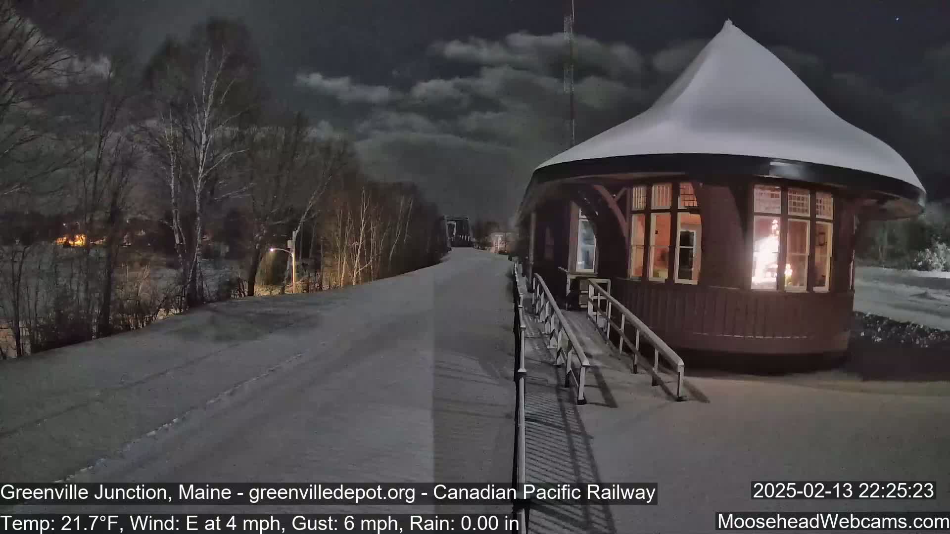 A snow-covered, round train station with a lit interior is seen at night, with a snow-covered road leading away from it under a cloudy sky.