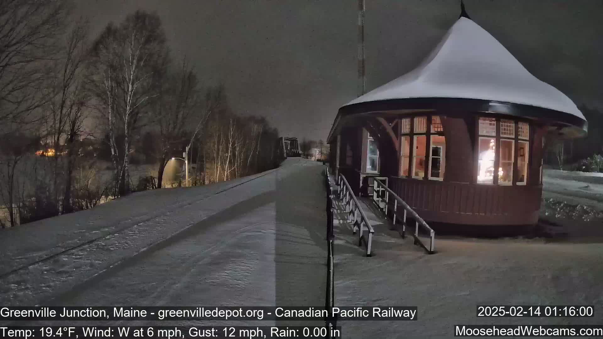 A snow-covered train depot with lights on inside sits next to a snow-covered railway track under a dark, nighttime sky.