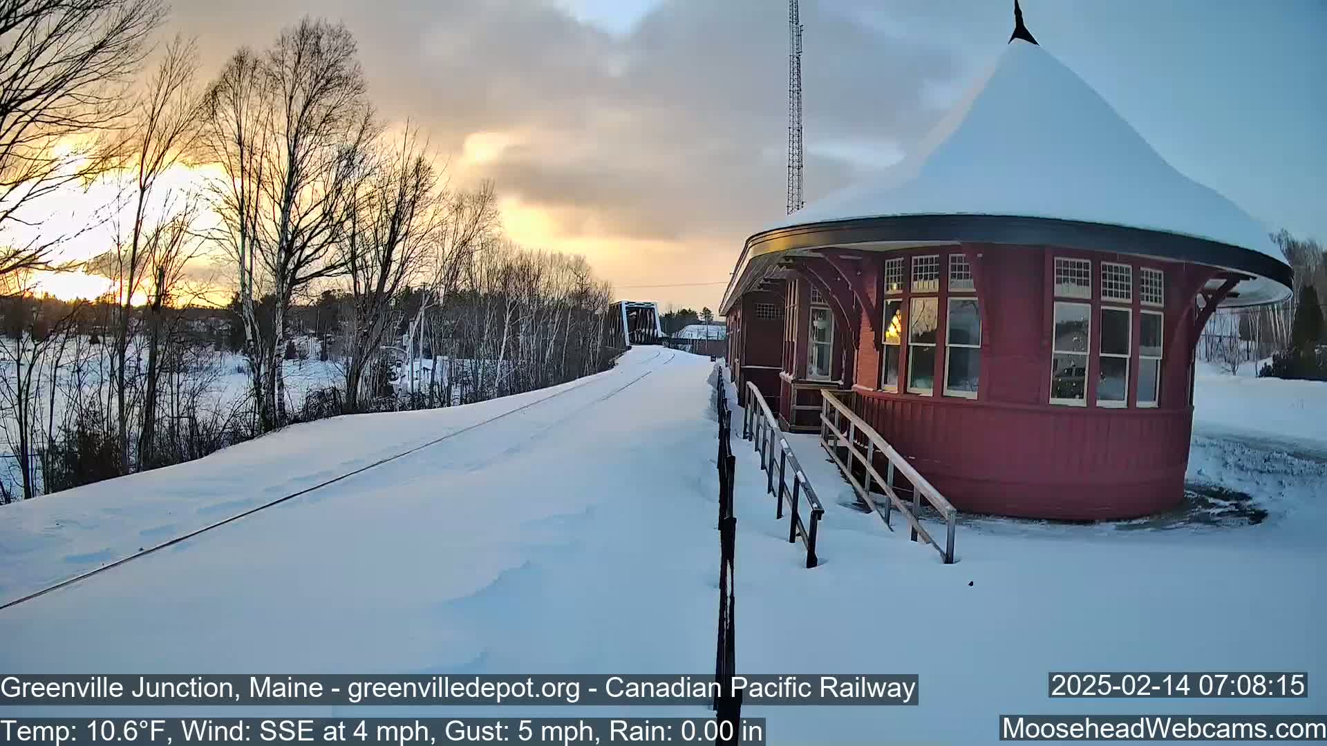 A snow-covered train platform with a red, round building is visible during a sunset under cloudy skies, with a train approaching in the distance.