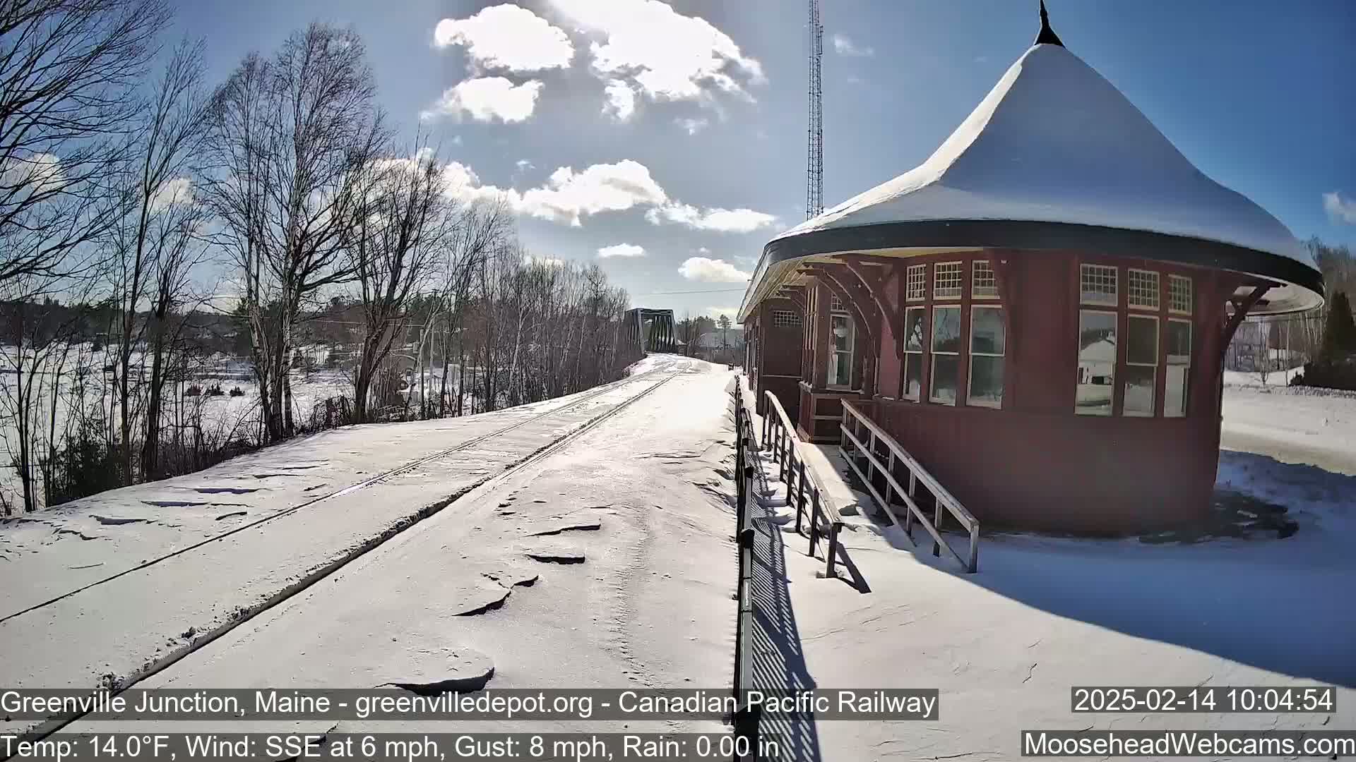 A snow-covered railway track leads to a red, snow-topped train station under a partly sunny sky.